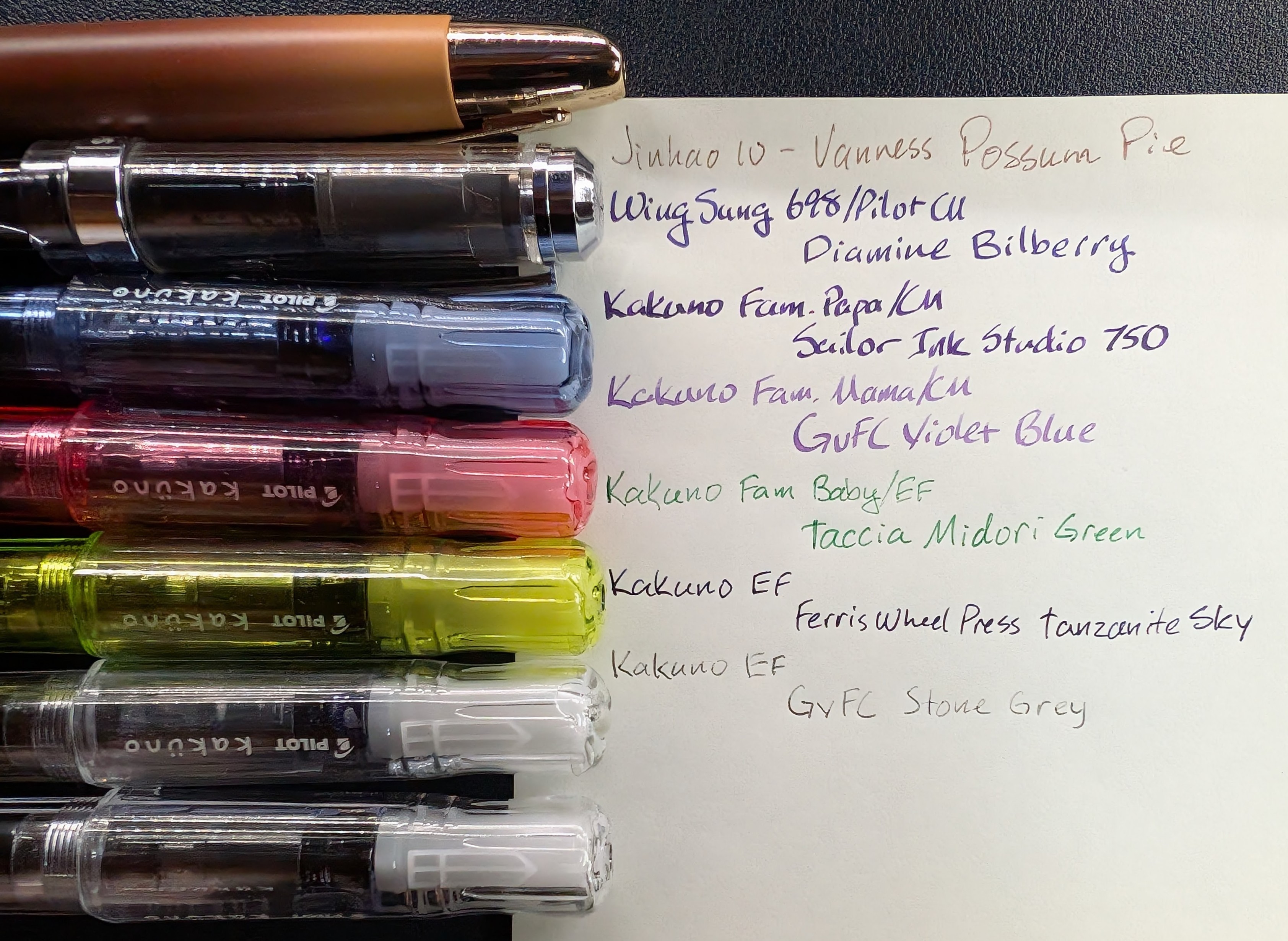 Photo of seven fountain pens next to a writing sample which includes the pen name and ink name. From top to bottom they are: - "Jinhao 10 - Vanness Possum Pie" (Light brown ink) - "WingSung 698/Pilot CM / Diamine Bilberry" (Dark purple ink) - "Kakuno Fam. Papa/CM / Sailor Ink Studio 750" (Dark blurple ink) - "Kakuno Fam. Mama/CM / GvFC Violet Blue" (Lavender Ink) - "Kakuno Fam. Baby/EF / Taccia Midori Green" (Bright green ink) - "Kakuno EF / Ferris Wheel Press Tanzanite Sky" (Dark blurple ink) - "Kakuno EF / GvFC Stone Grey" (Light gray ink)