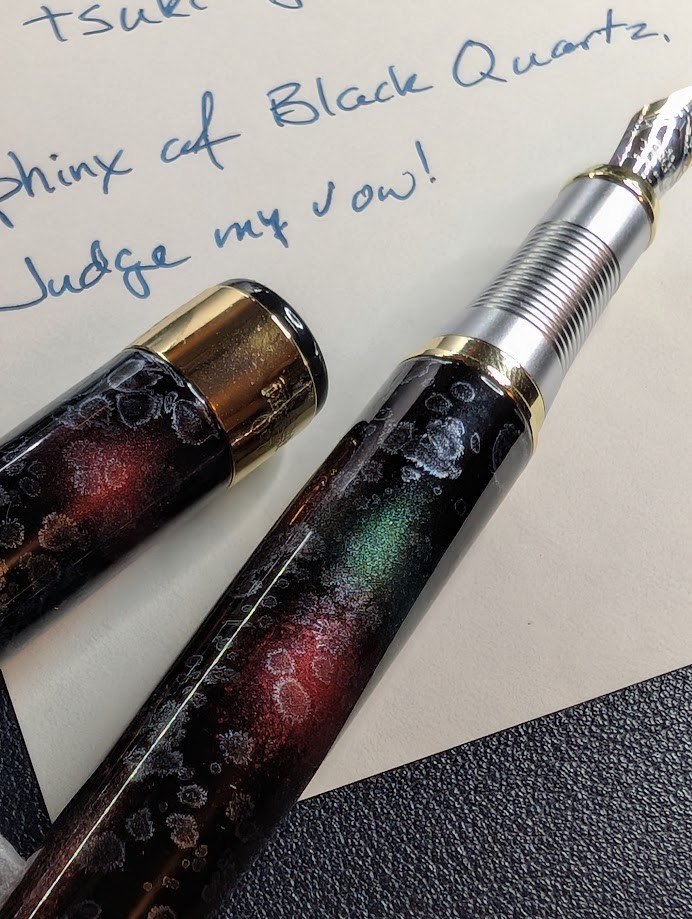 Photo of a writing sample and fountain pen. The pen is uncapped and the cap is laying next to the pen. This photo is focused primarily on the pen. The writing sample is in ink that is dark blue with a hint of green. It is only partially visible but says "Sphinx of Black Quartz, Judge my vow!". The pen is a dark base color, blue nearly black. There are curcular bursts of red and green on the pen. There are also whiteish areas that look like various shapes of droplets with thicker outlines. The section on the pen is silver and ribbed. The nib is two-tone silver and gold plating. The accents on the cap and around the section are gold.