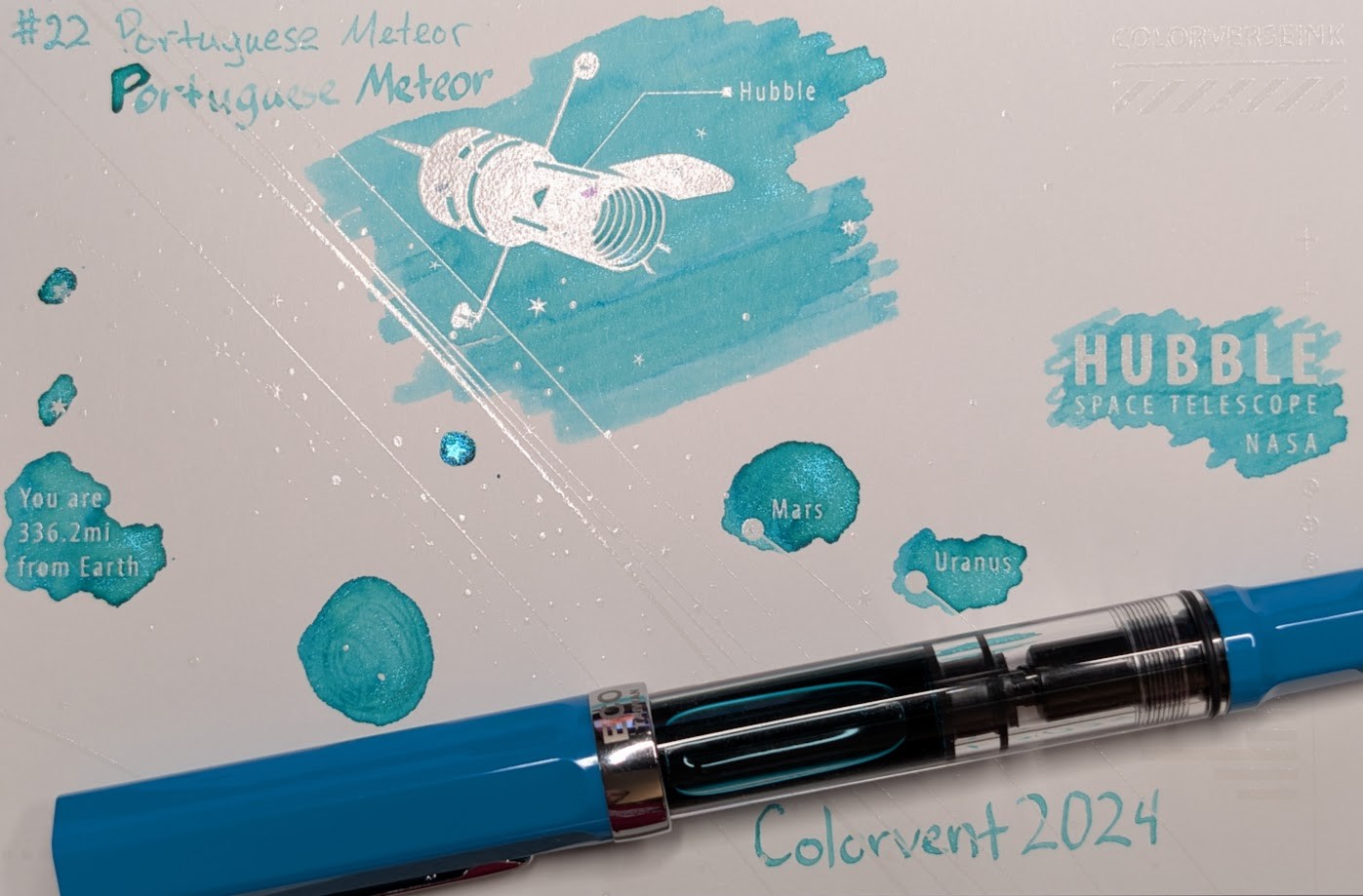 Photo of a "Colorverse Colorspace Size C" swatch card, type 2 which is based on the Hubble space telescope. The ink on the card is a bright aqua/turquoise color with low shading and cyan shimmer. Upper left is text reading "#22 Portuguese Meteor" written in an EF nib and then a stub nib. Just to the left side of the middle at the top is a large illustration of the Hubble space telescope in negative space among swabbed ink. In the middle right side is text "Hubble Space Telescope NASA" in negative space among scribbled ink. In the middle left is text reading "You are 336.2mi from Earth" in negative space among ink scribbles. In the middle lower left is a blob of swirled ink drawn by a glass dip pen. Near the center are "Mars" and "Uranus" with circles representing those planets, in more swabbed ink. Lower right is text "Colorvent 2024". Laying across the card is a TWSBI Eco fountain pen, with Cerulean Blue cap/tail and silver accents, 1.1 stub nib.