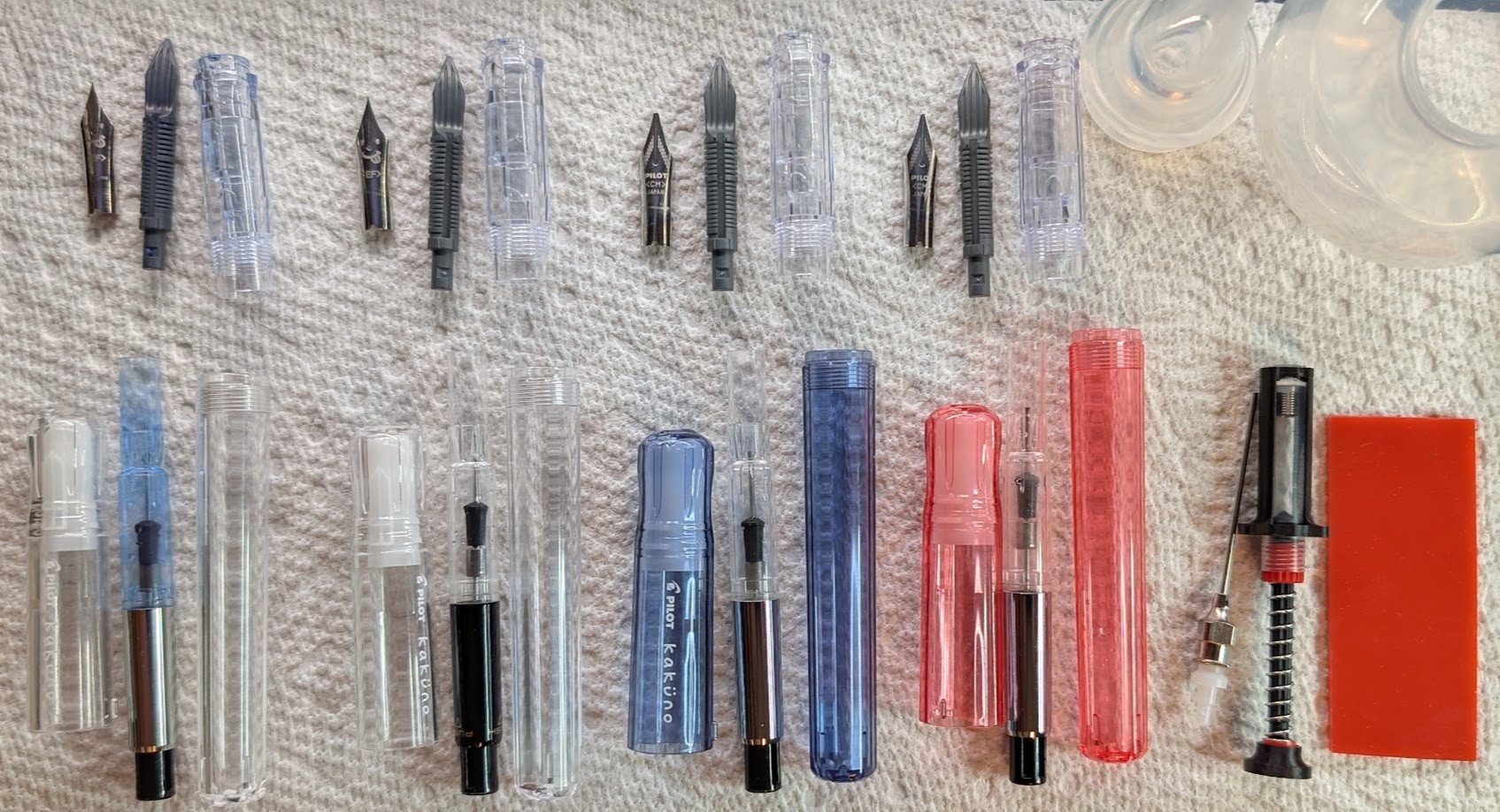 Photo of four disassembled fountain pens on a paper towel. Next to the pens is a disassembled clear bulb syringe, a spring-loaded converter syringe, and a bit of rubber. The four fountain pens are all Pilot Kakunos, two are clear with EF nibs. One is transparent blue, and the last one is transparent red. Each pen is separated out completely: Cap, body, converter, section, feed, and nib all separate and grouped.