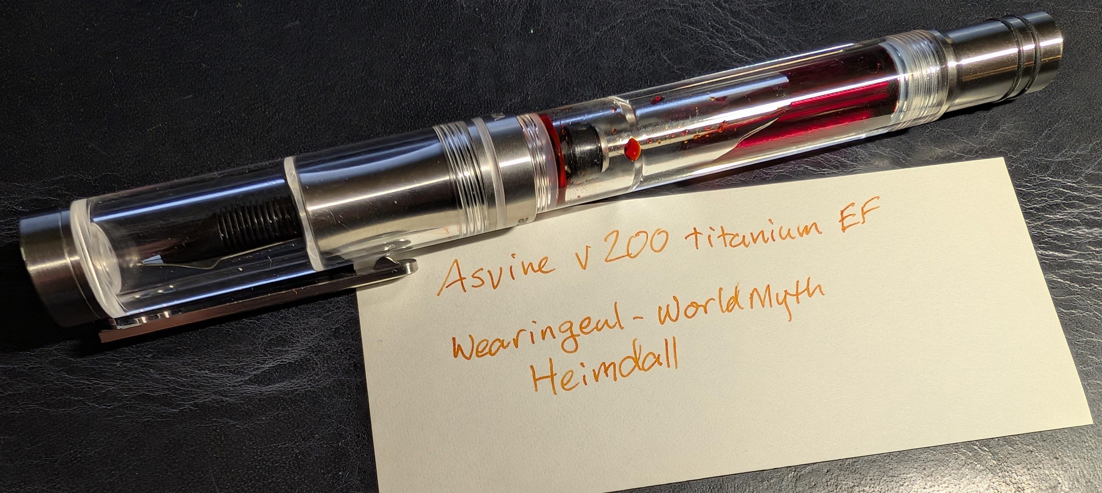 Photo of a fountain pen next to a writing sample written in orange ink. The pen is a clear vacuum filler with titanium accents. Orange ink is visible inside. The writing sample reads "Asvine V200 Titanium EF / Wearingeul - World Myth / Heimdall"