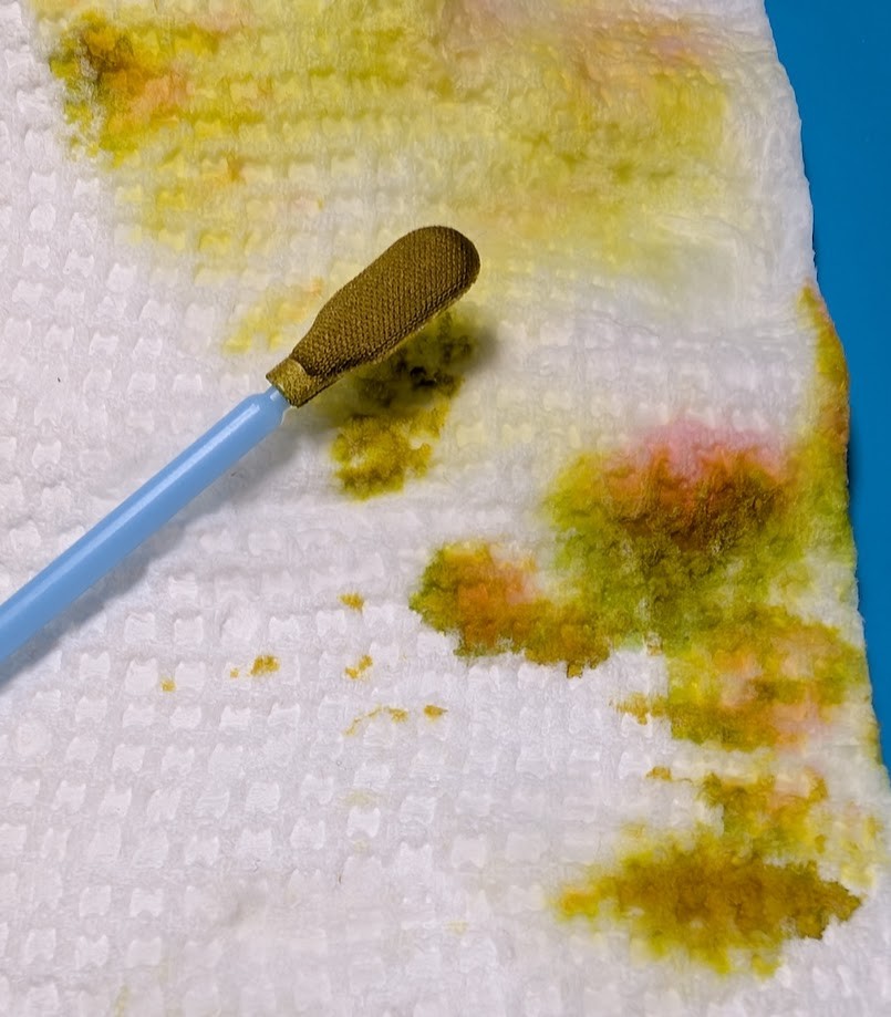 Photo of a paper towel and lint-free swab. Both have ink on them. The ink on the swab is very dark brown, maybe a little yellow, and a little green. The ink on the paper towel is mostly yellow but also a sickly green and some red/pinkish areas.