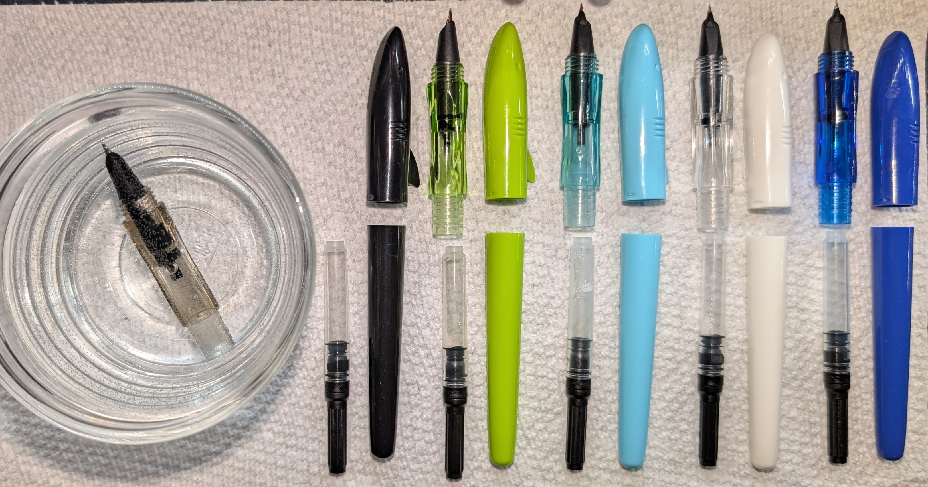Photo of five disassembled fountain pens on a paper towel. Each pen is separated into four parts: Nib+Section, Converter, Cap, and Body. Each is arranged in a similar manner, except for the pen on the left has its nib section soaking in a small glass ramekin full of water. The pens are all Jinhao Shark pens, which have a tapered body and a cap with a shark head design (dorsal fin, gills, mouth/eyes). From left to right the colors are: Black, Green, Light Blue, White, and Dark Blue.