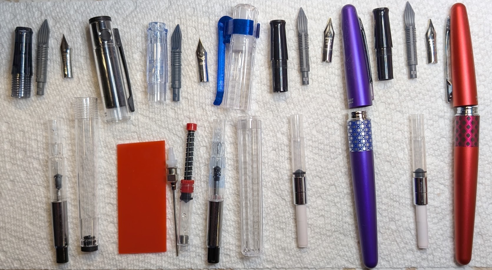Photo of four disassembled fountain pens on a paper towel, along with a bit of rubber and a spring-loaded converter syringe. The pens are, from left to right: - Pilot Explorer/Lightive, clear with black trim, Fine nib, and CON-70 converter - Pilot Kakuno, clear with a blue 3D printed clip, EF nib, and CON-70 converter - Pilot Metropolitan Retro Pop, purple, CM nib, WingSung converter - Pilot Metropolitan Retro Pop, red, CM nib, WingSung converter