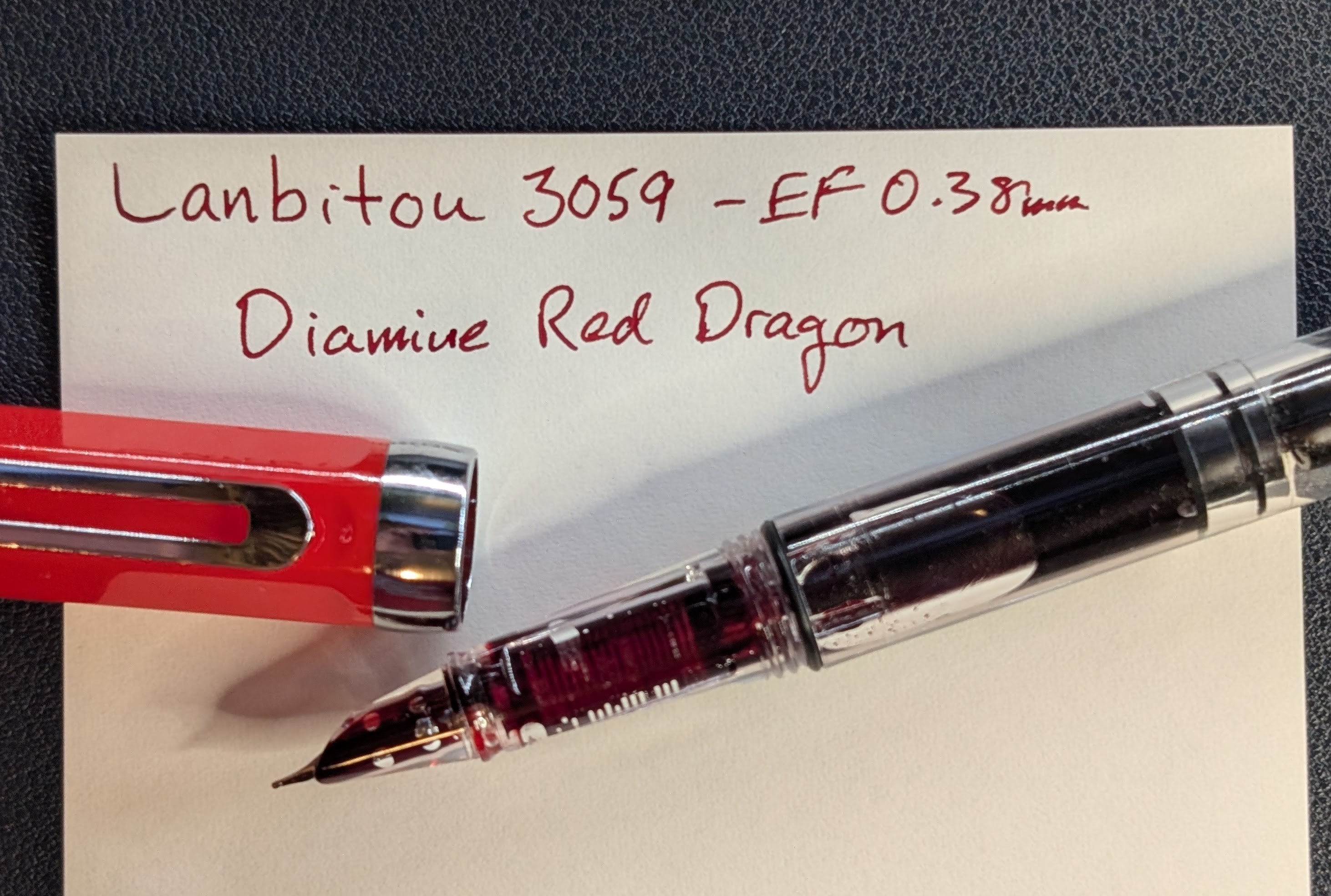 Top-down photo of an uncapped fountain pen made of transparent plastic, and a writing sample on paper. The cap is bright red with silver accents. The pen section is completely transparent, as is the feed inside. Both are saturated with red ink. The nib is hooded in transparent plastic and there is visible ink between the hood and the top of the nib. In is visible inside the body of the pen. The writing sample is in red ink and reads "Lanbitou 3059 - EF 0.38mm / Diamine Red Dragon"