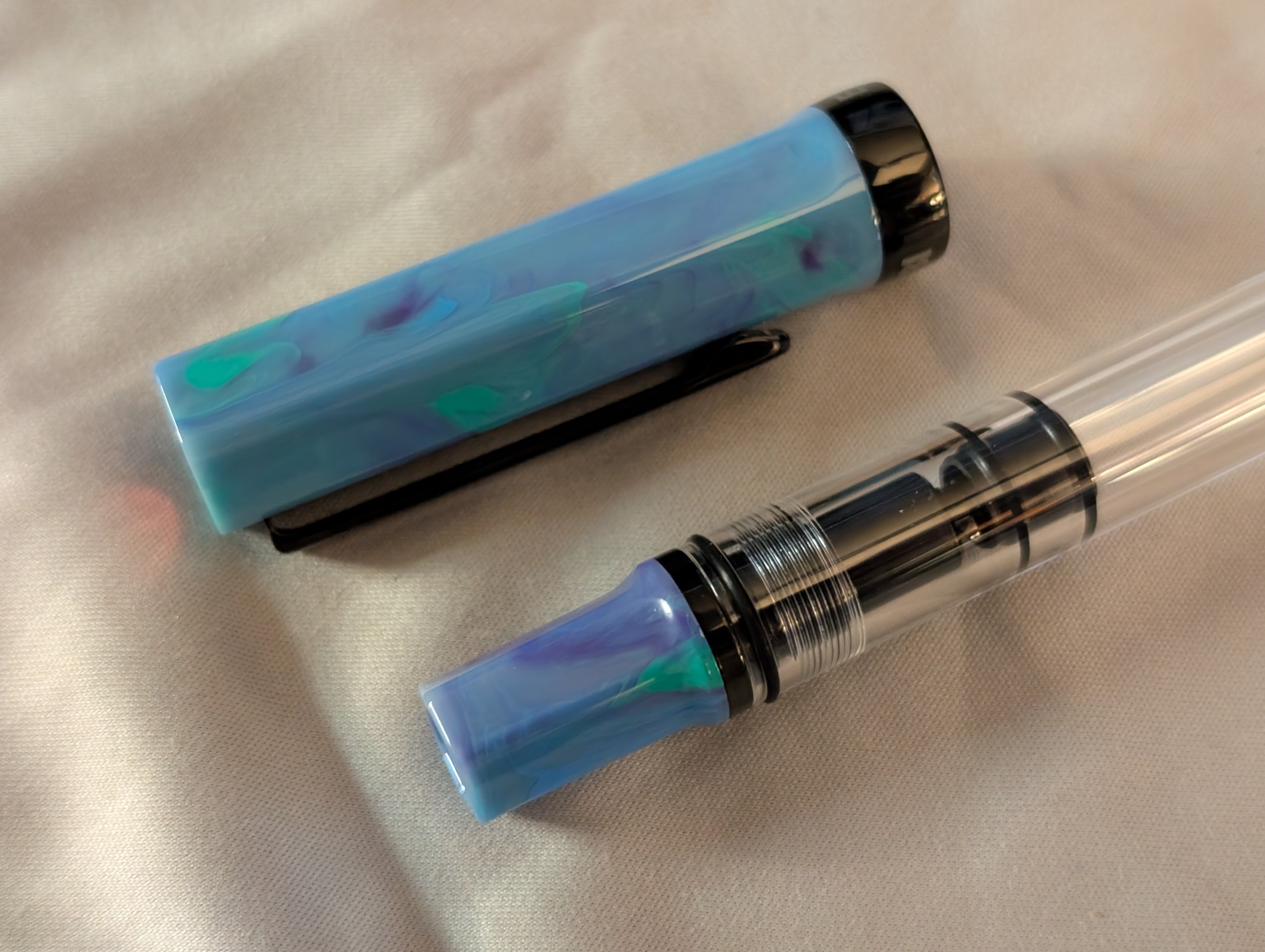 Photo of an uncapped fountain pen with the cap laying next to the pen. The tail end of the pen is situated next to the cap. The pen cap and piston knob are a swirled mix of blue with streaks of purple and green.