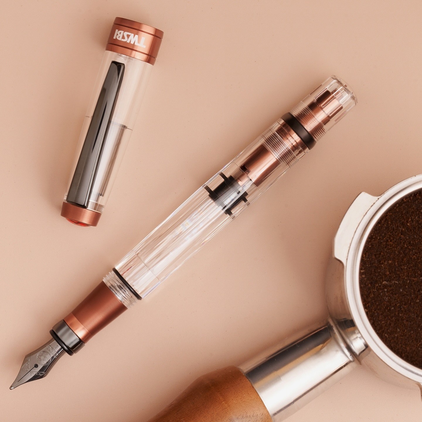 Photo of a fountain pen next to an espresso machine portafilter full of compacted coffee grounds. The background is very light brown. The pen has a clear body with brown metal about the same color as coffee around the grip section and inside the piston knob, with dark "onyx" trim and nib. In this photo the brown appears a bit "cooler" and less ruddy than the other photos.