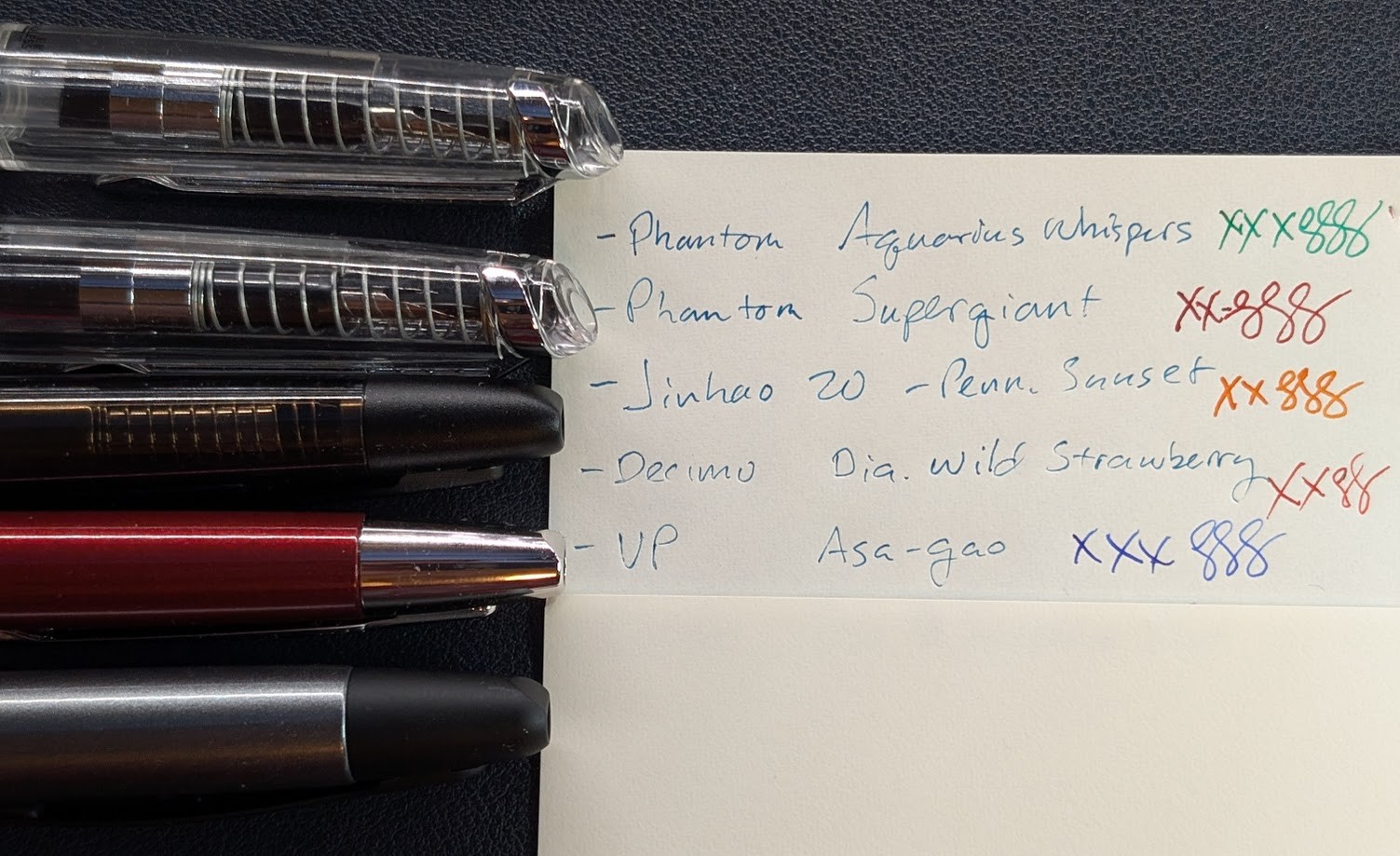 Five fountain pens in a row, only the tips are visible, next to a piece of paper. The pens each correspond to a line on the paper. - Endless Phantom (clear demonstrator) - Paper says "Phantom Aquarius Whispers" followed by "XXX" and a few curlicues in green ink. - Endless Phantom (clear demonstrator) - Paper says "Phantom Supergiant" followed by "XX" and a few curlicues in dark red ink. - Jinhao 20 in dark brown transparent plastic with matte black trim - Paper says "Jinhao 20 - Penn. Sunset" followed by "XX" and a few curlicues in orange ink. - Pilot Vanishing Point Decimo in burgundy with silver trim - Paper says "Decimo Dia. Wild Strawberry" followed by "XX" and a few curlicues in bright red ink. - Pilot Vanishing Point in gun metal with matte black trim - Paper says "VP Asa-gao" followed by "XXX" and a few curlicues in blue ink.