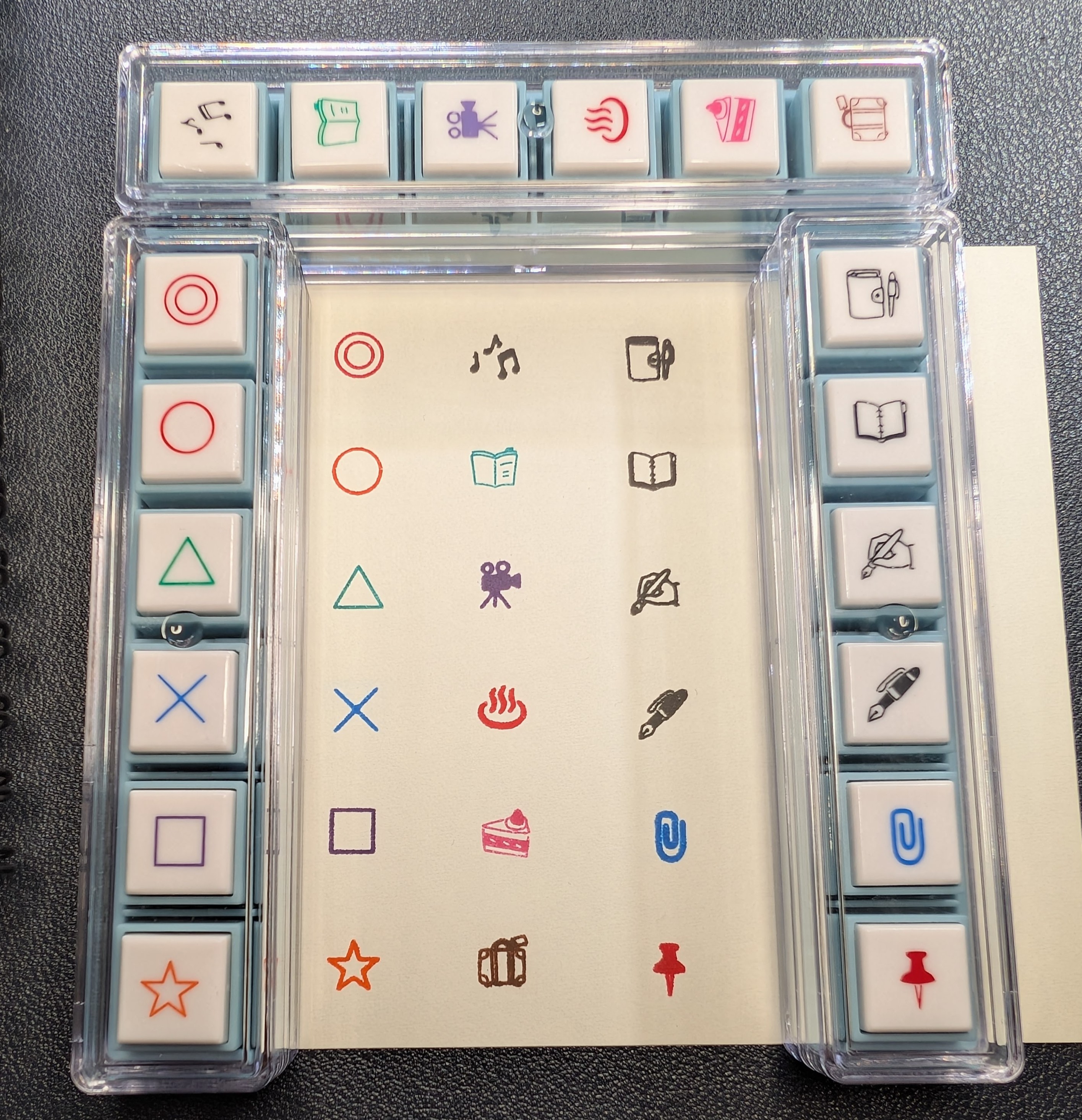 Photo of three six-stamp sets sitting on a piece of paper showing the marks the stamps made. To the left: A red target, orange circle, green triangle, blue X, purple square, and red star. Across the top: Black music notes, green book (cover side), purple movie camera, red plate with steam rising, pink piece of cake, brown suitcase To the right: A black closed notebook with pen in a loop, an black open blank notebook, a black pen in a hand writing, a black fountain pen, a blue paper clip, and a red pushpin/thumbtack