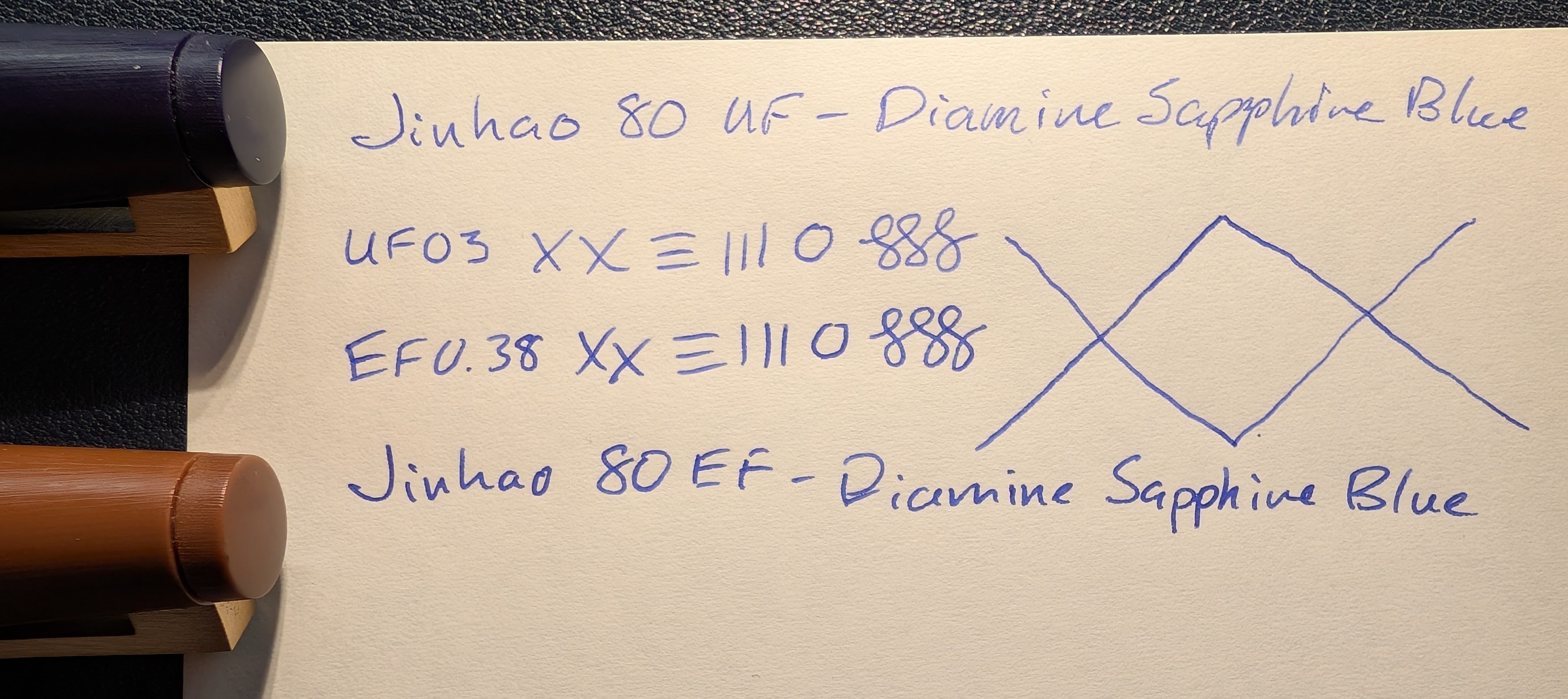 Photo of a writing sample and lines drawn on paper with two pens next to the paper. The pens only have the end of the caps visible. Top is blue, bottom is brown, both with gold clips. The writing sample reads "Jinhao 80 UF - Diamine Sapphire Blue" across the top with "UF03 XX , three vertical and horizontal lines, a circle, and three curlicues. Under that it is similar but "EF0.38" and the lines are slightly thicker. The bottom reads "Jinhao 80 EF - Diamine Sapphire Blue". To the right of the center are two large intersecting lines. A thinner one starting at the top that goes down and back up. Then a slightly thicker one that starts at the bottom and goes up and back down. Together they look like "XX"