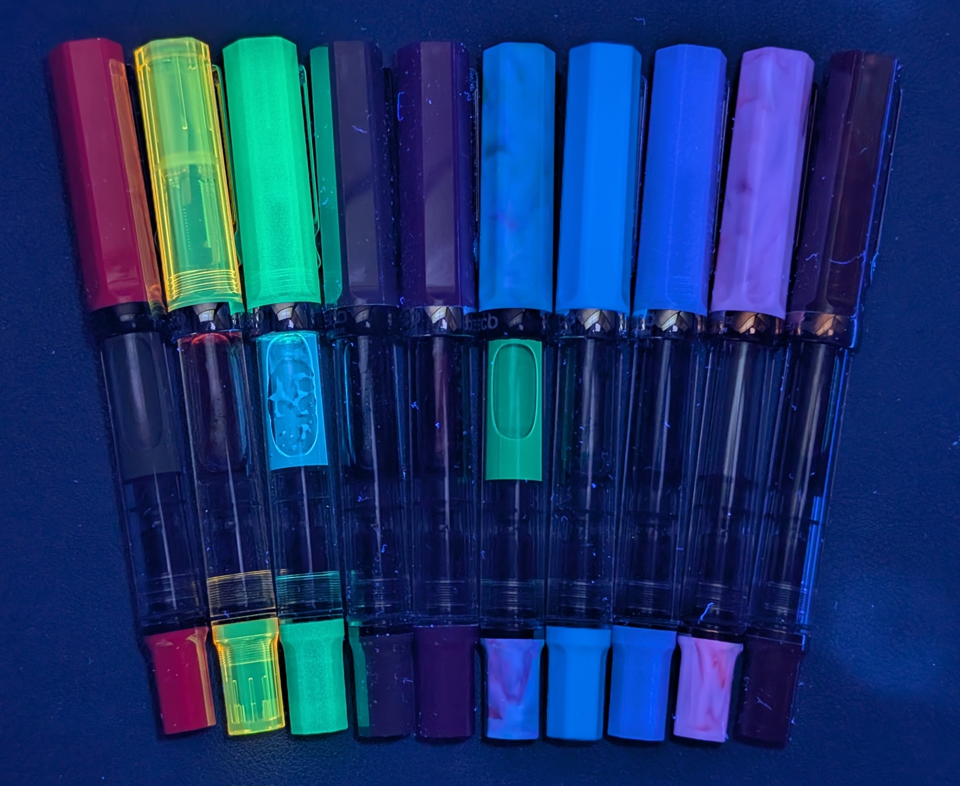 Photo of several TWSBI ECO fountain pens under UV light. From left to right they are: Plum (glowing a wee bit) Transparent Yellow (glowing brightly) Glow Green (glowing very brightly, ink inside is glowing blue) Slate Blue (Not Glowing) Dark Sapphire (glowing maybe a teeny tiny bit) Cosmos Blue (glowing a lot, ink inside is glowing green) Cerulean Blue (glowing a lot) Glow Purple (glowing brightly) Fluorite (glowing brightly) Root Beer (not glowing)