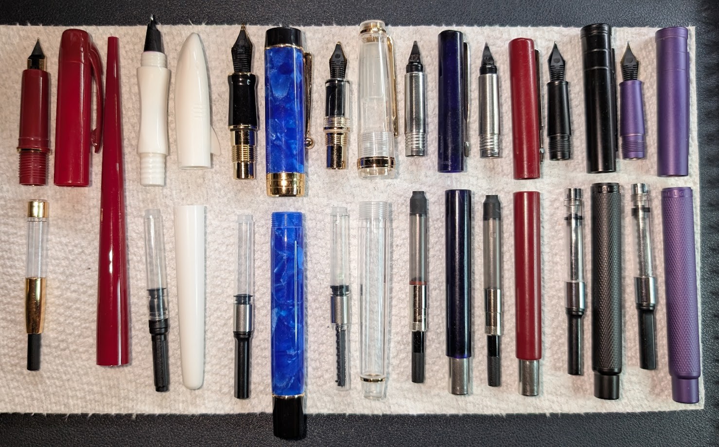Photo of 8 disassembled fountain pens on a paper towel. Each is separated into four pieces and arranged the same way: Nib+Section, Converter, Cap, Body. From left to right the pens are: - Platinum Desk Pen (Red, EF) - Jinhao Shark (White/White, EF) - Jinhao 100/Century (Haibao Blue, Fude) - Jinhao 82 (Clear, Fude) - Parker Vector (Blue, FI) - Parker Vector (Red, M) - Hongdian Black Forest (Black, EF) - Hongdian Purple Forest (Purple, EF)