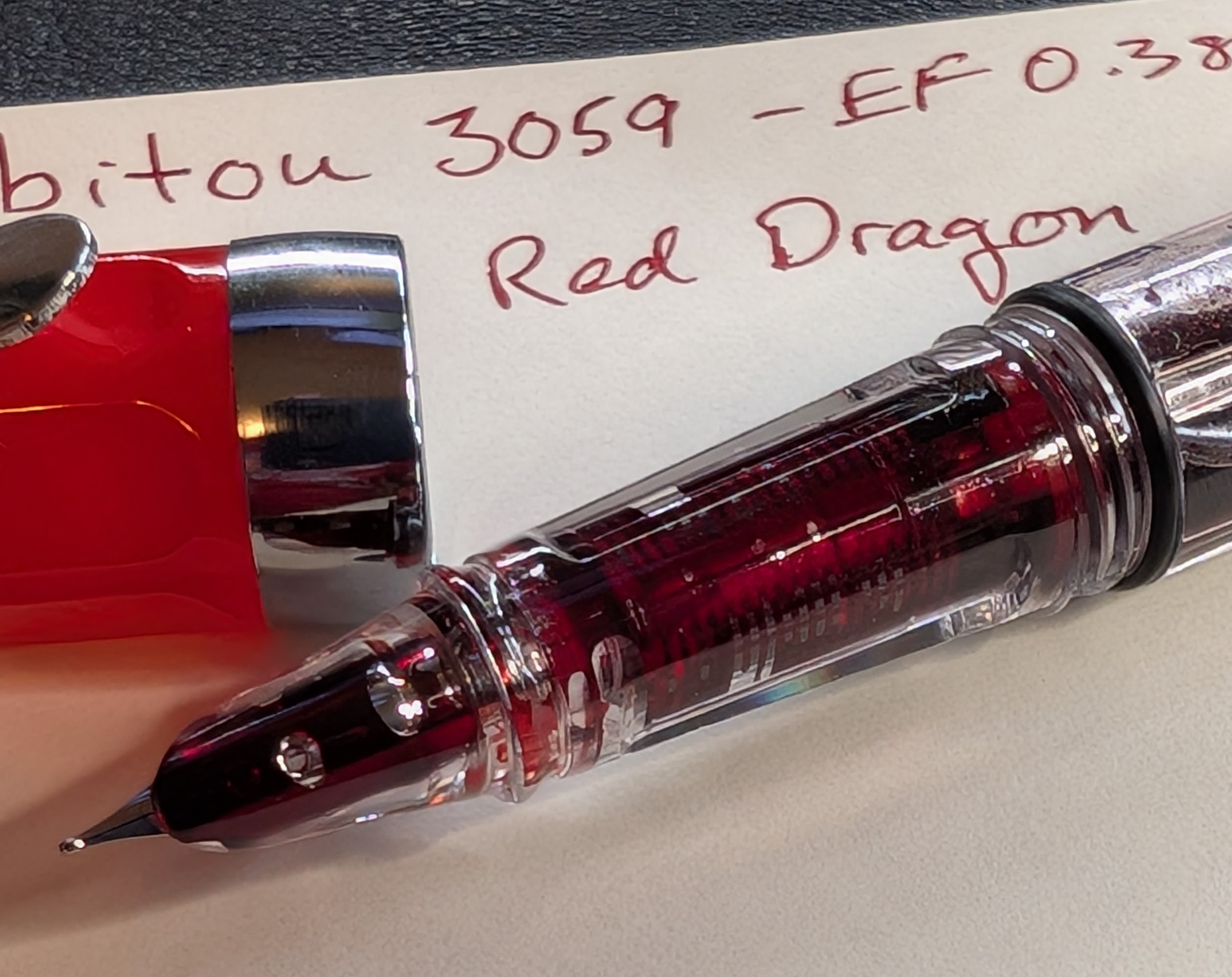 Close-up oblique angle photo of an uncapped fountain pen and writing sample. Focus is on the pen nib, the writing is not all visible, but is written in red ink. The cap is bright red with silver accents. The pen section is completely transparent, as is the feed inside. Both are saturated with red ink. The nib is hooded in transparent plastic and there is visible ink between the hood and the top of the nib.
