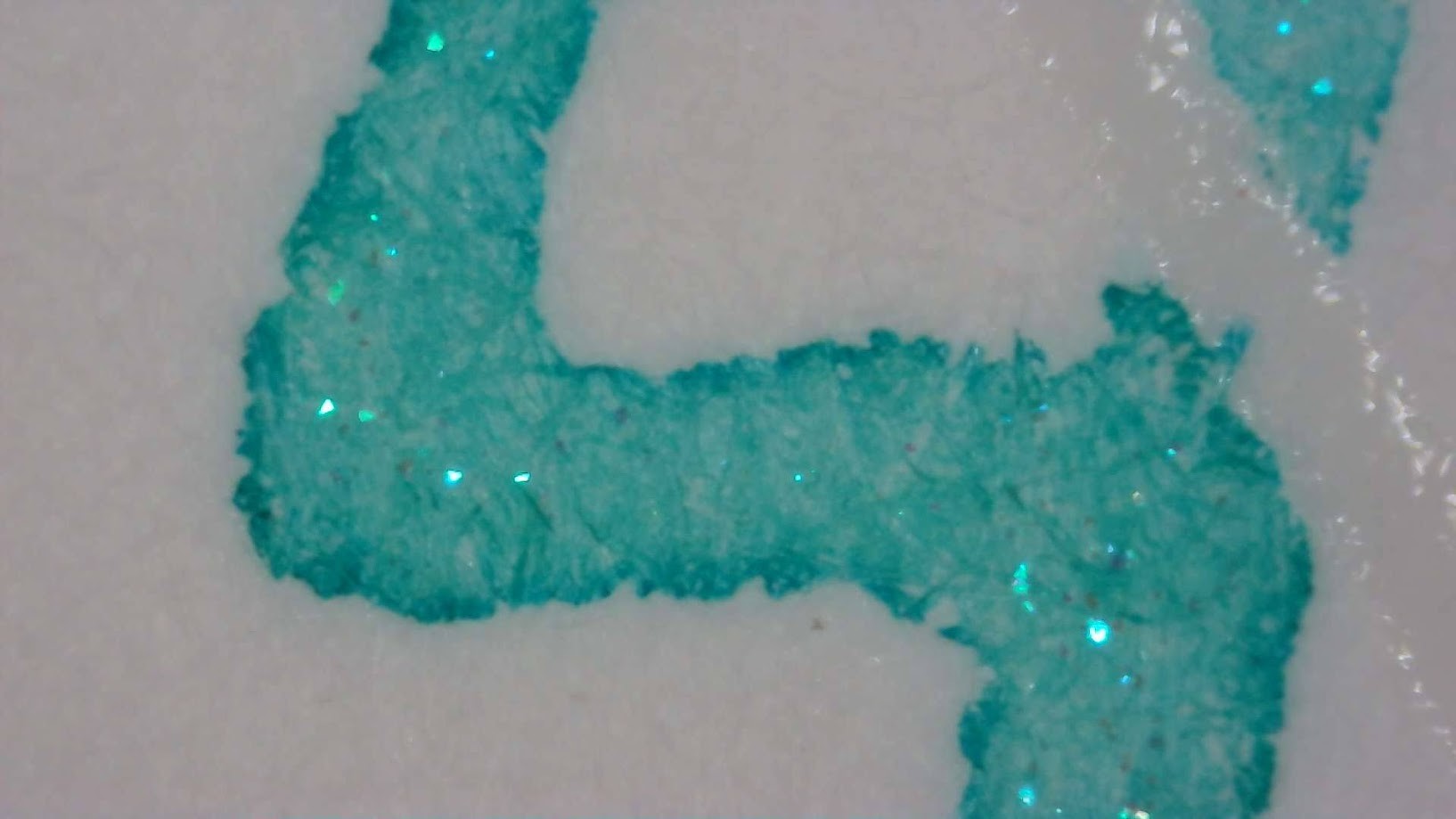 Photo of ink writing under magnification. Paper fibers are visible. The ink is a bright aqua/turquoise color. The edges of the lines are slightly darker. There are cyan shimmer flakes occasionally inside the lines.