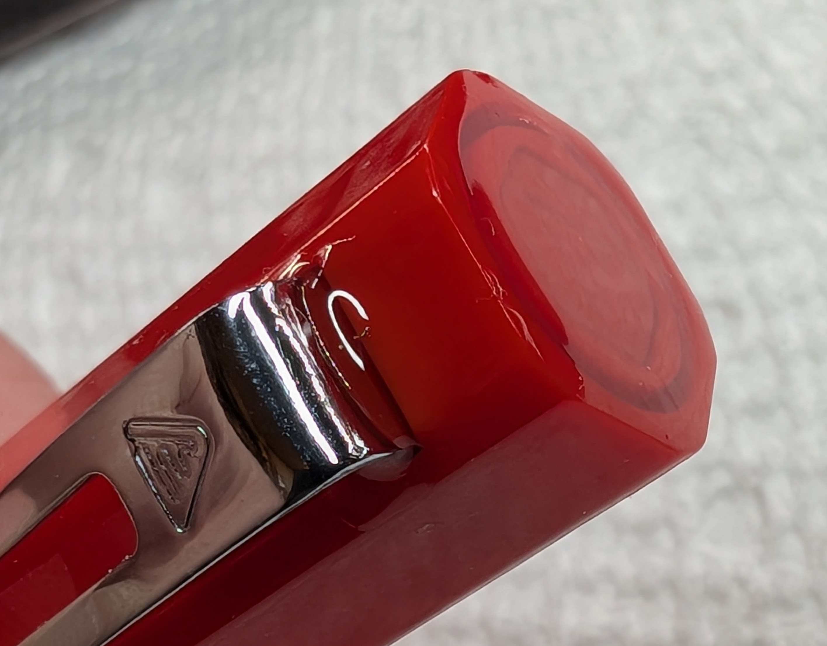 Photo of the end of a red hexagonal fountain pen cap. The finial is visible as well as the clip. The entire finial surface is shiny as it has been completely encased by resin. The base of the clip goes into a hole on the side of the cap and where the two parts meet is also shiny as it's full of resin.