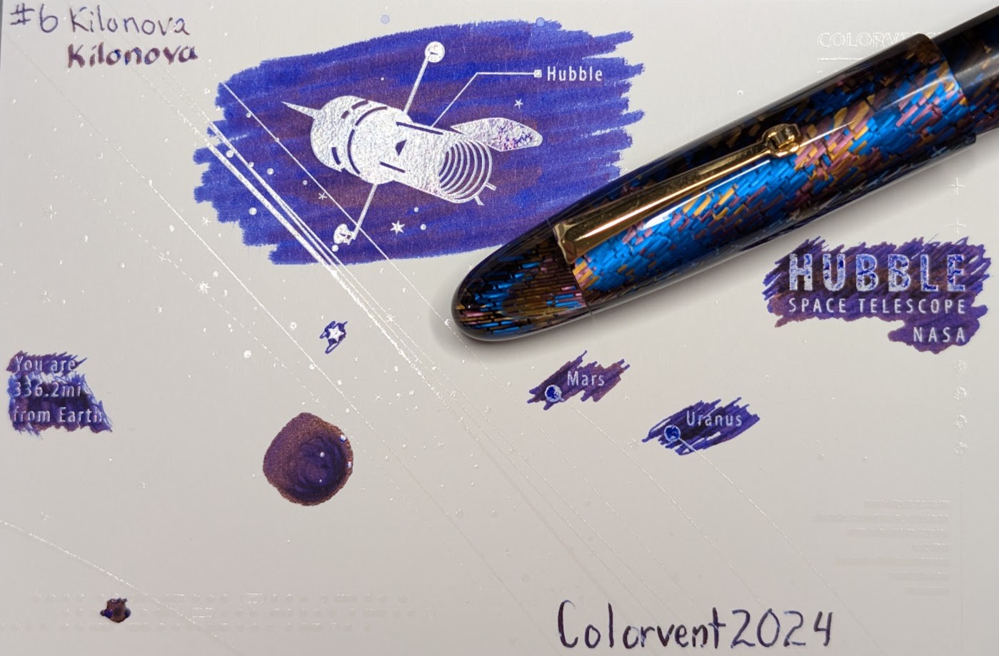 Photo of a "Colorverse Colorspace Size C" swatch card, type 2 which is based on the Hubble space telescope. The ink on the card is a blue-purple "blurple" ink with reflective red/purple sheen where ink has pooled. Upper left is text reading "#6 Kilonova" written in an EF nib and then a stub nib. Just to the right side of the middle at the top is a large illustration of the Hubble space telescope in negative space among swabbed ink. In the middle right side is text "Hubble Space Telescope NASA" in negative space among scribbled ink. In the middle left is text reading "You are 336.2mi from Earth" in negative space among ink scribbles. In the middle lower left is a blob of swirled ink drawn by a glass dip pen. Near the center are "Mars" and "Uranus" with circles representing those planets, in more ink. Lower right is text "Colorvent 2024". Laying across the card is a Vazir Meteor shower fountain pen, which is blue with streaks of blue, purple, and yellow stacked celluloid.