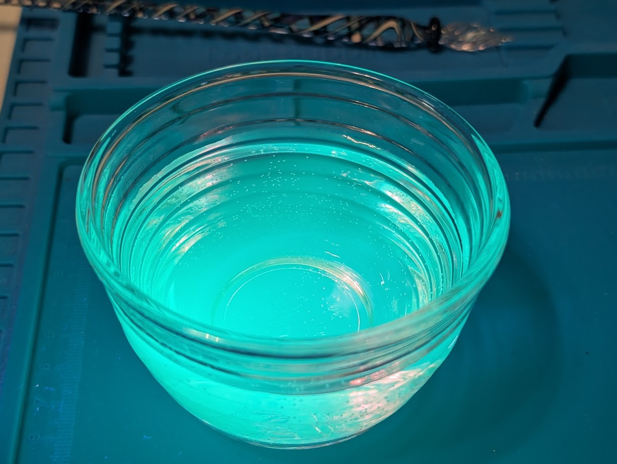 Photo of a glass ramekin filled with liquid, it is eerily glowing light green under UV light and looks like it might taste like key lime or maybe radioactive pear. The ramekin is sitting on a blue silicone mat, and a glass dip pen is visible in the background. Alas, the liquid is actually water with some very diluted fluorescent fountain pen ink from cleaning a dip pen, and would probably taste gross and likely make me sick.
