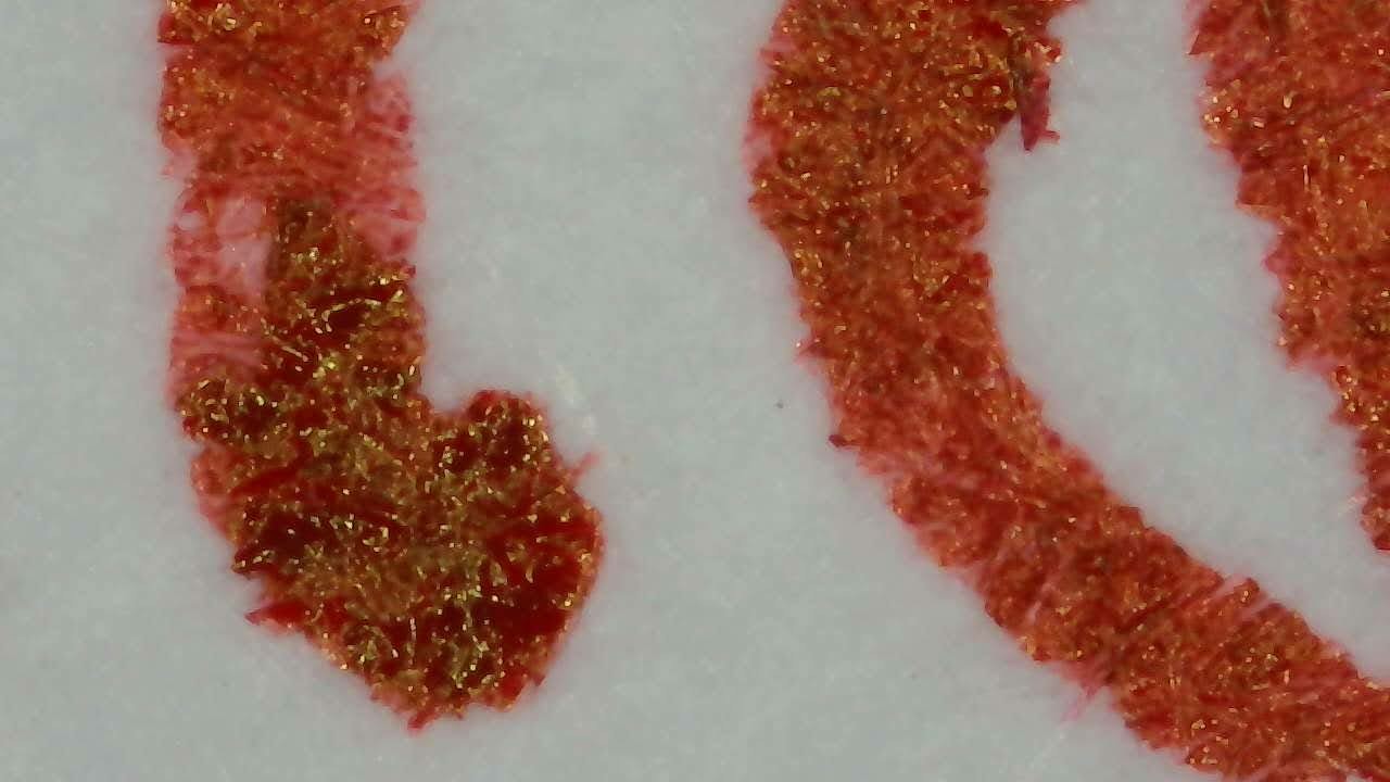 Photo of ink on paper taken with a microscope camera. Paper fibers are visible throughout. There are several curved lines of red ink. In some areas of the red ink, little shiny flecks of gold color are visible.