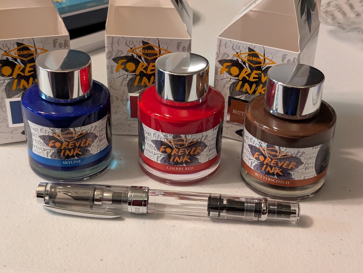 Photo of three fountain pen ink bottles in front of their open boxes. They are all "Diamine Forever" inks. From left to right they are: Skyline, a light blue. Cherry Red, a bright bold red. Butterscotch, a medium brown. In front of the bottles lies a TWSBI Diamond 580 fountain pen with clear plastic body, cap, and no colored accents. The clip and ring are silver.