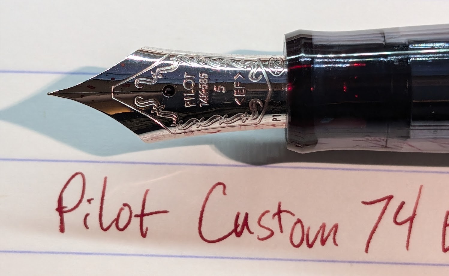Photo of a fountain pen nib and writing sample on a pad of paper. There is text engraved on the nib "Pilot 14k-585 5 <EF> P1118" There is an engraved outline around the nib with some vaguely S-shaped curlicue type designs inside the outer line. The writing sample is in dark red ink and reads "Pilot Custom 74"