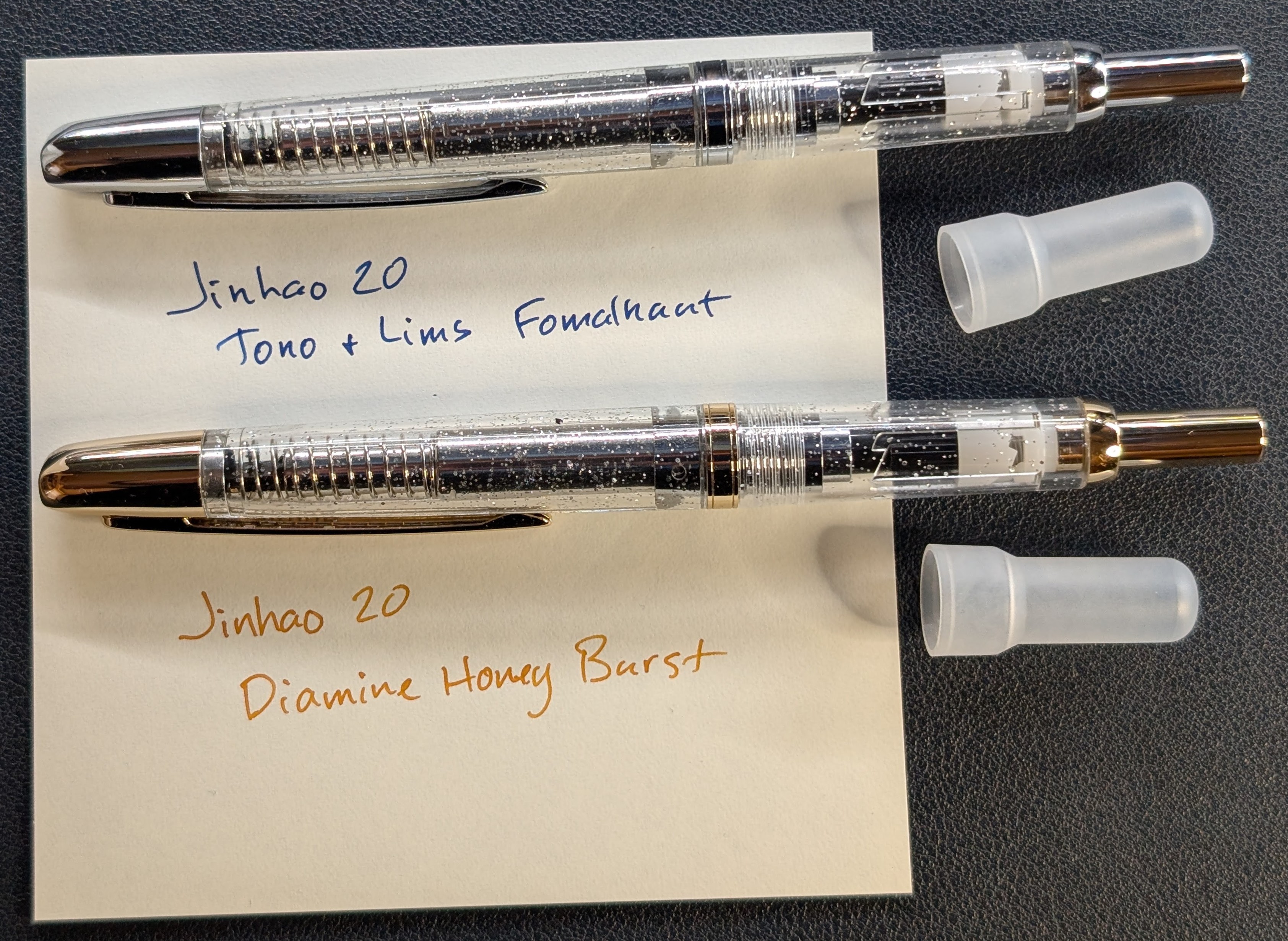 Photo of two capless fountain pens laying across a piece of paper with writing. A protective cap that covers the tail button is laying next to each one. The top pen is transparent with silver trim. The writing below the pen is in dark blue ink and reads "Jinhao 20, Tono + Lims Fomalhaut" The bottom pen is transparent with gold trim The writing below the pen is in a honey-colored dark orange and reads "Jinhao 20, Diamine Honey Burst"