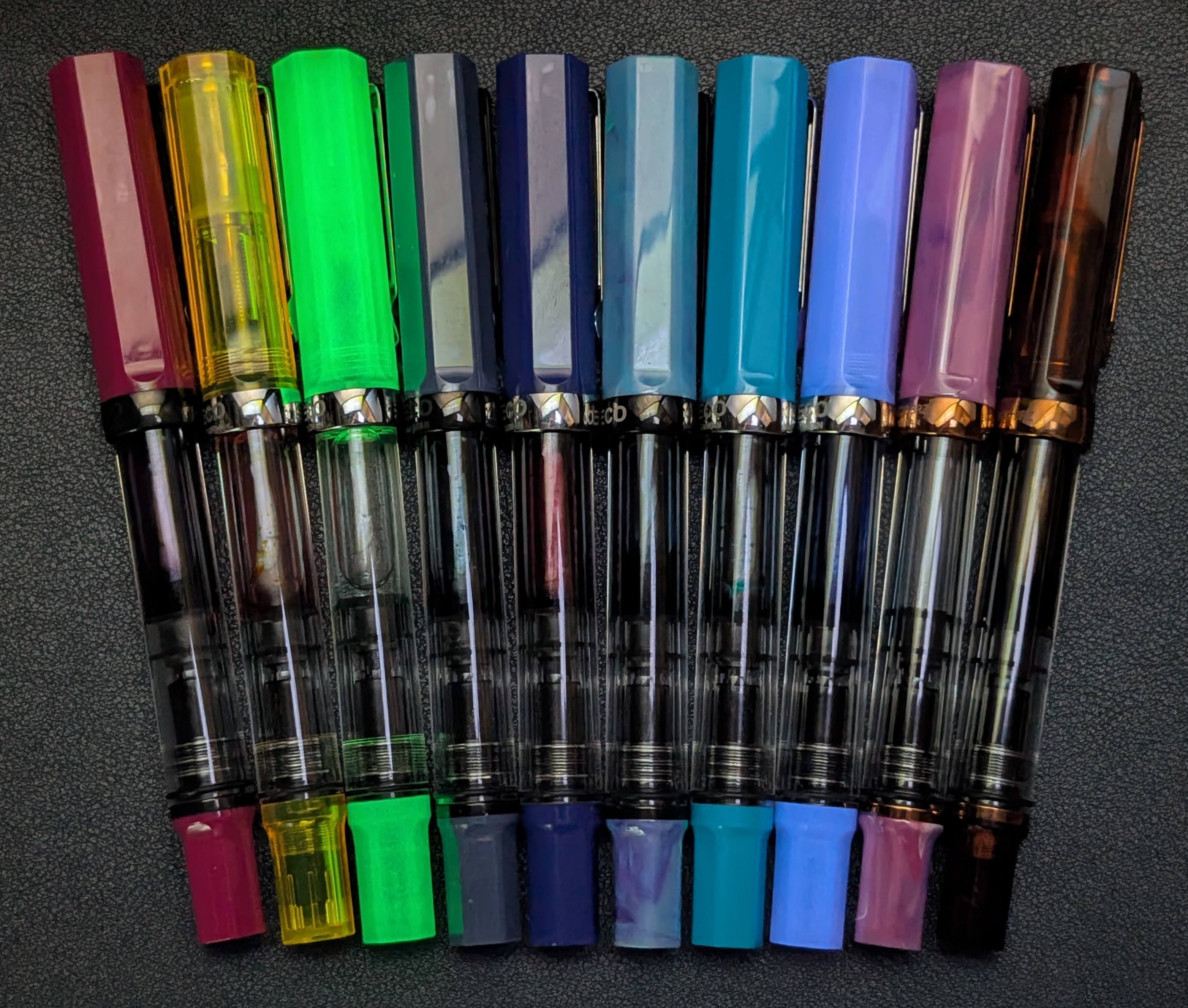 Photo of several TWSBI ECO fountain pens under UV light. From left to right they are: Plum Transparent Yellow Glow Green (glowing very brightly) Slate Blue Dark Sapphire Cosmos Blue Cerulean Blue Glow Purple (glowing brightly) Fluorite Root Beer