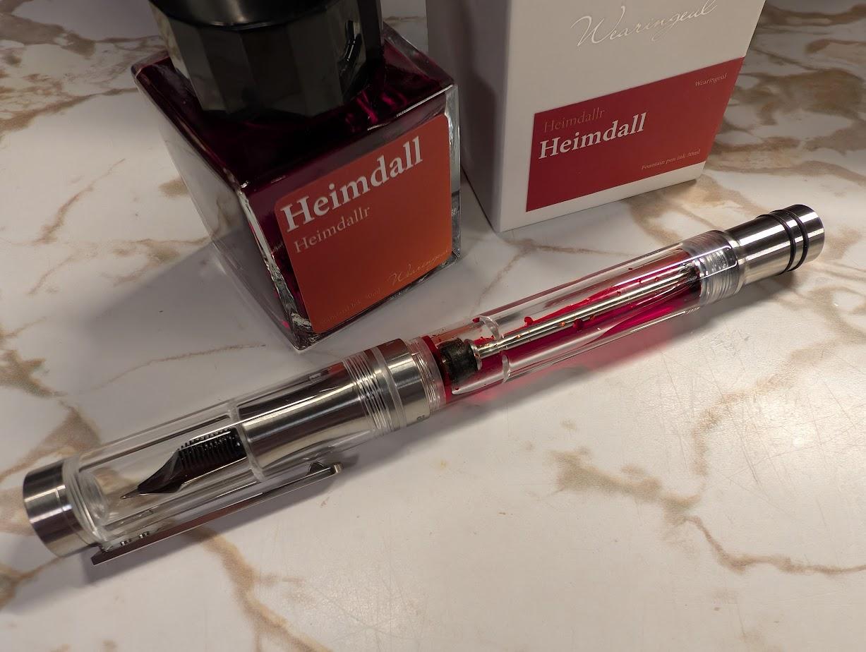 Photo of a fountain pen next to a bottle of orange ink and its box. The pen is a clear vacuum filler with titanium accents. Orange ink is visible inside. The label on the ink and box both read "Heimdall" and have the brand name "Wearingeul"