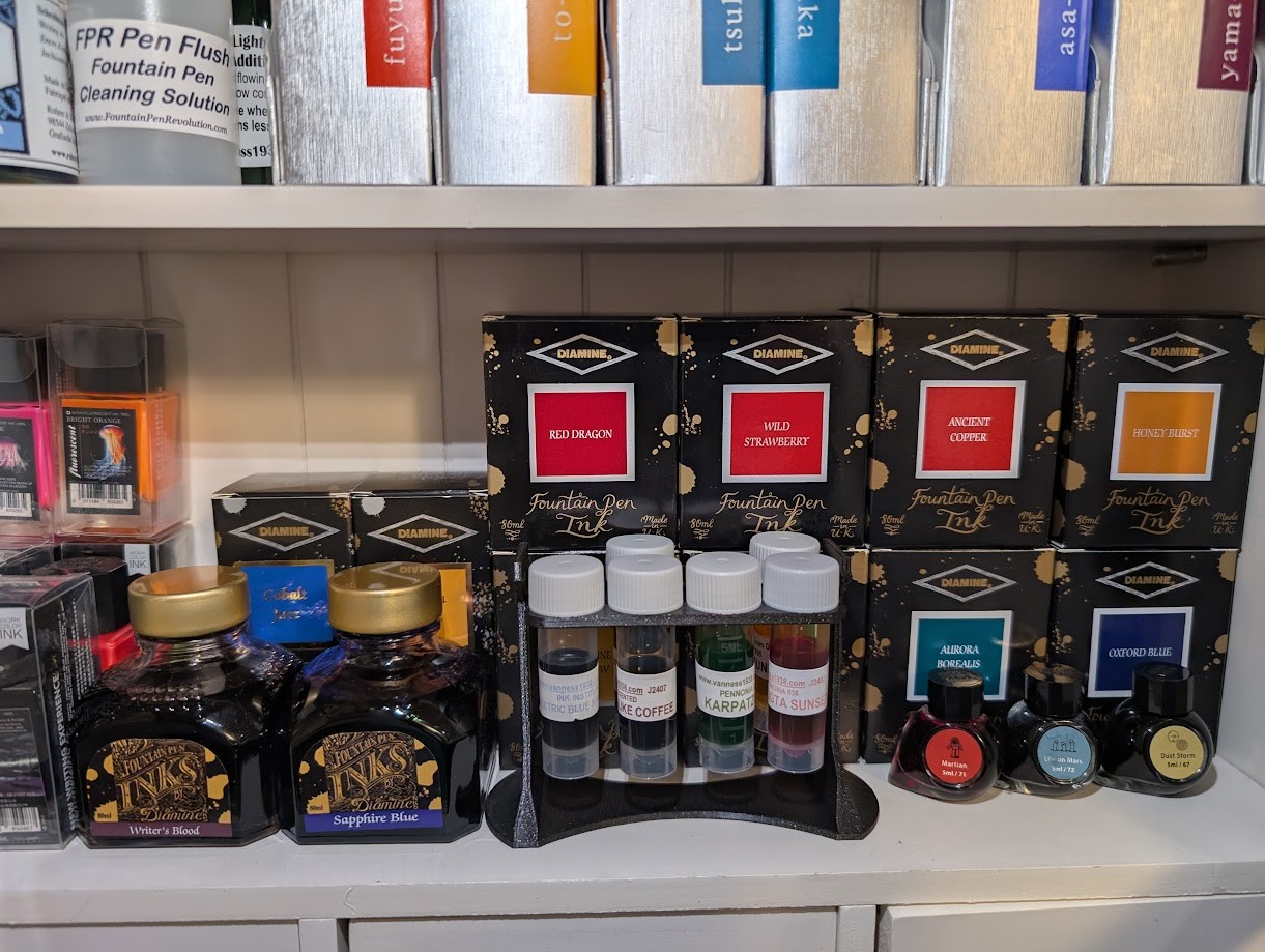 Photo of a 3D printed sample vial rack on a small shelf among numerous other ink bottles. There are sample six vials in the rack and two empty spaces.