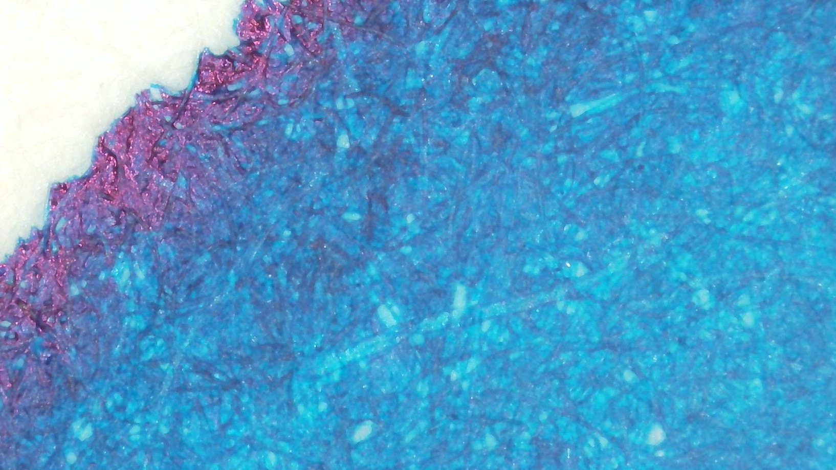 Photo of ink blob on paper under magnification. Paper fibers are visible. The ink is mostly medium blue-green but toward the edge it gets darker and the edge itself has a reflective reddish tone.