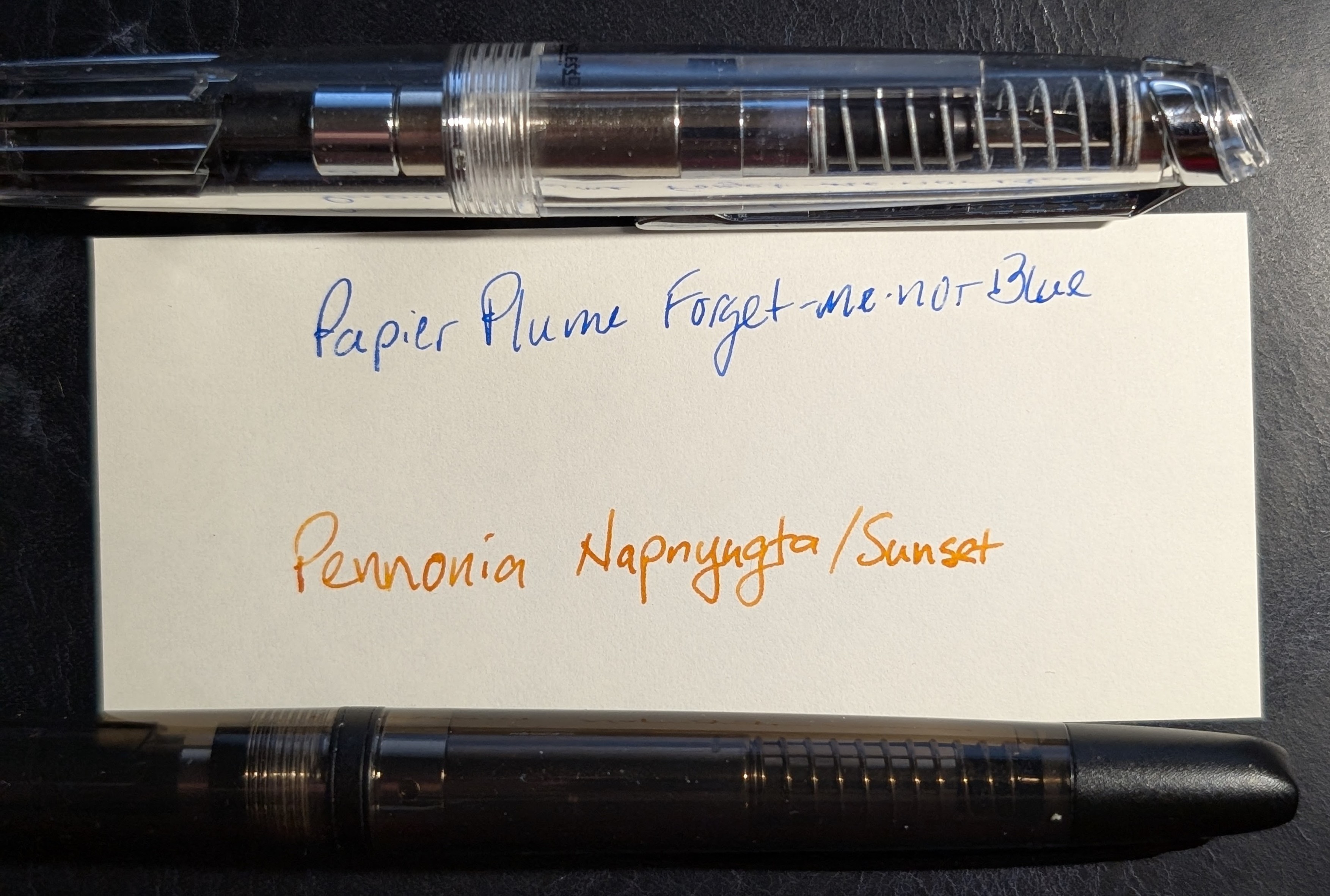 Photo of two fountain pens laying next to a strip of paper with writing. Across the top is an Endless Phantom capless fountain pen demonstrator (clear) with silver accents and beneath that is writing in blue ink "Paiper Plume Forget-me-not Blue" Next is orange writing "Pennonia Napnyugta/Sunset" and beneath that is a Jinhao 20 with semi-transparent black plastic body and matte black accents.
