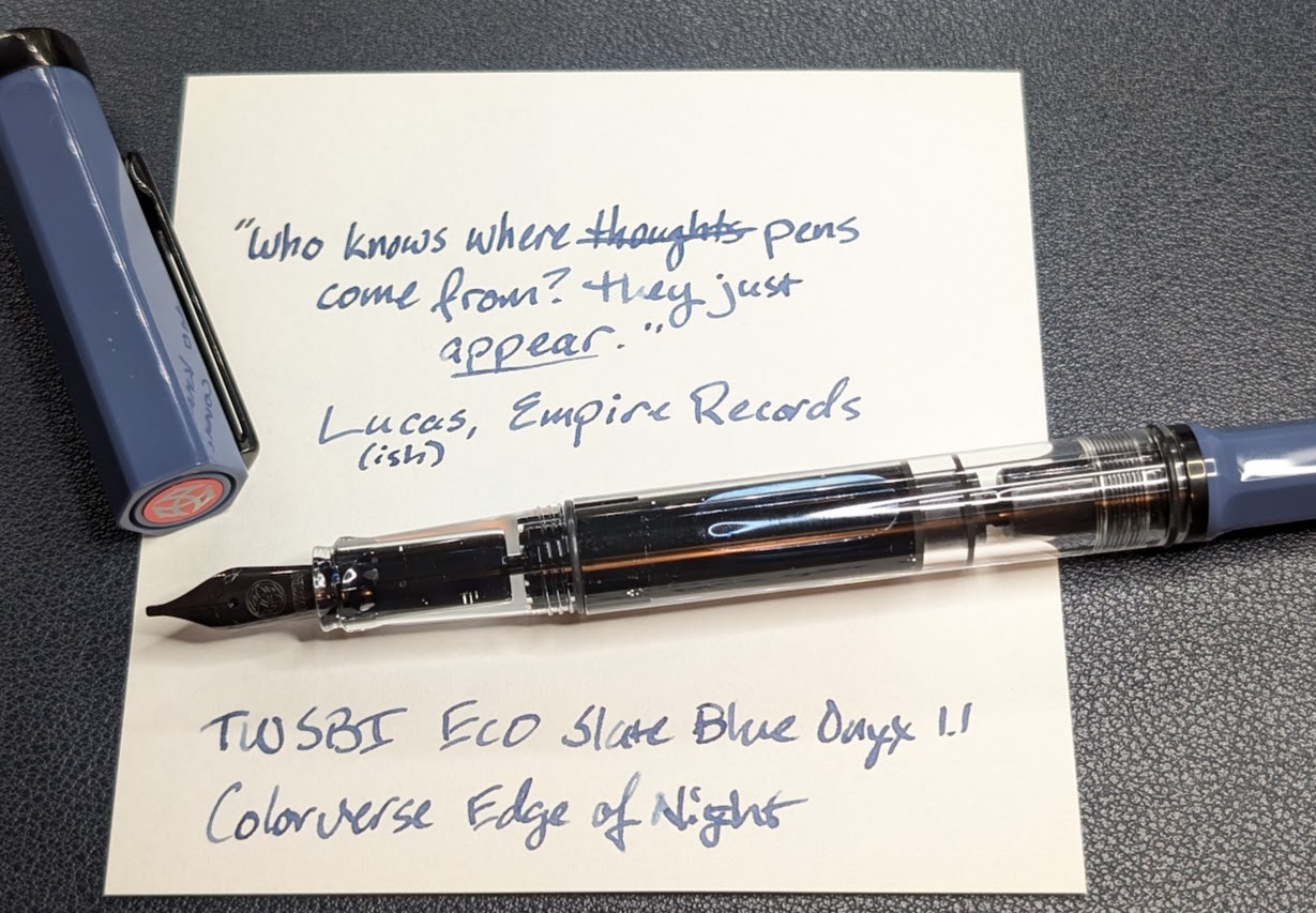 Photo of a desk containing a fountain pen and writing sample on a piece of note paper. The photo is taken from the top-down at the slight angle downward from the front. The fountain pen is uncapped, the cap a dark blue-gray color and is laying off to the left at an angle with its finial pointing toward the bottom center of the photo. The pen has a black nib, clear section and body, and a dark blue-gray tail. It is a piston filling pen with blue ink visible inside. The writing sample reads: "Who knows where thoughts come from? They just appear." Thoughts is crossed out and replaced by "pens". The quote is attributed to "Lucas (ish), Empire Records" There is more writing under the pen: "TWSBI ECO Slate Blue Onyx 1.1", "Colorverse Edge of Night"