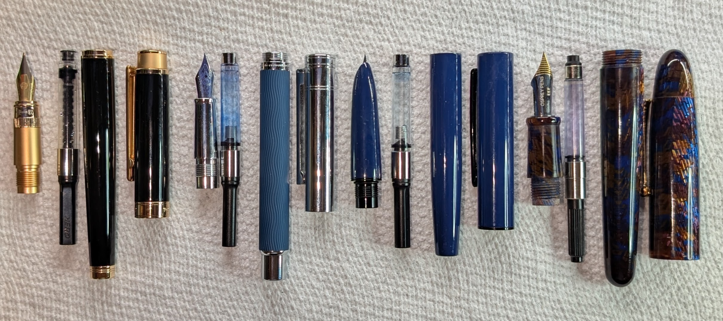Photo of four disassembled fountain pens arranged in a line with the top of each piece aligned. Each pen is separated into four parts: Nib+Section, converter, body, cap. From left to right they are: - Hero 1086 adjustable flex nib pen, black with gold accents - Hongdian 1843 Voyager, blue and silver, the converter is stained blue. - Hongdian C1 hooded nib in blue - Vazir Meteor Shower in blue