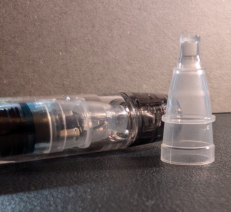 Tip of a Platinum Preppy fountain pen with its cap on, laying on a desk next to a roughly conical-shaped bit of plastic which is standing up. The conical plastic piece is identical to the inner cap visible through the clear plastic lid on the pen next to it.