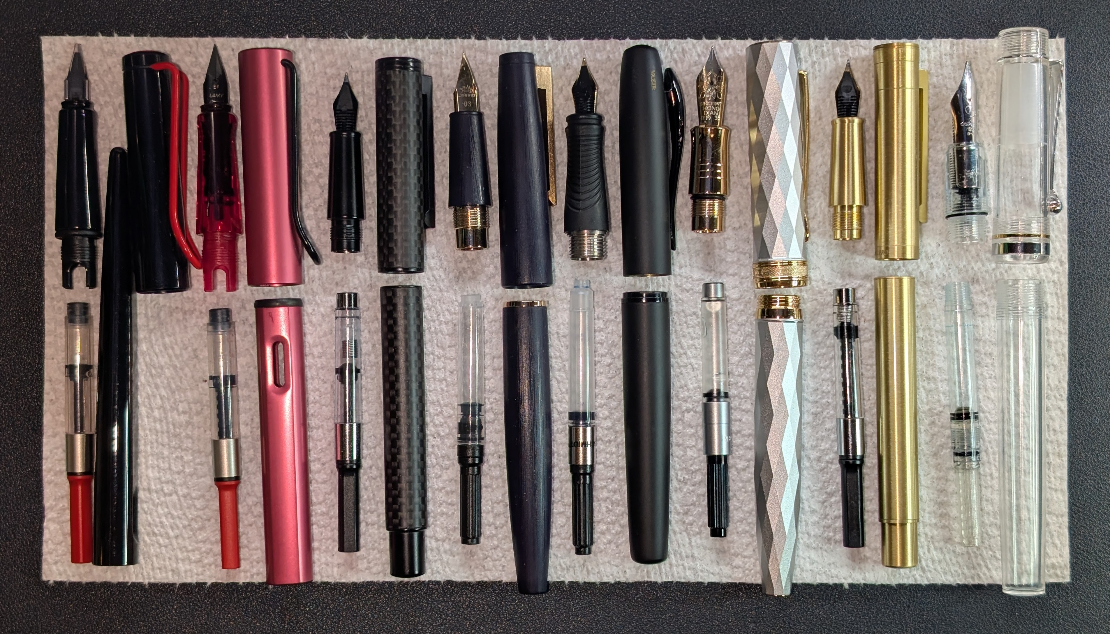 Photo of 8 disassembled fountain pens on a paper towel. Each pen is separated into four parts: Nib+Section, Converter, Body, and Cap. They are all lined up with parts arranged nearly the same way. From left to right the pens are: Lamy Joy (black with red trim, 1.9mm Stub), Lamy Al-Star (Fiery Red, EF), Hongdian Black Forest Pro Carbon Fiber (EF), Jinhao 80 (Blue, UF03), Vazir Onyx (Black), Hongdian N11 (Silver Lozenge, EF), Hongdian 1861 (Raw Brass, Long Blade), Osprey Madison (Clear, Architect Broad)