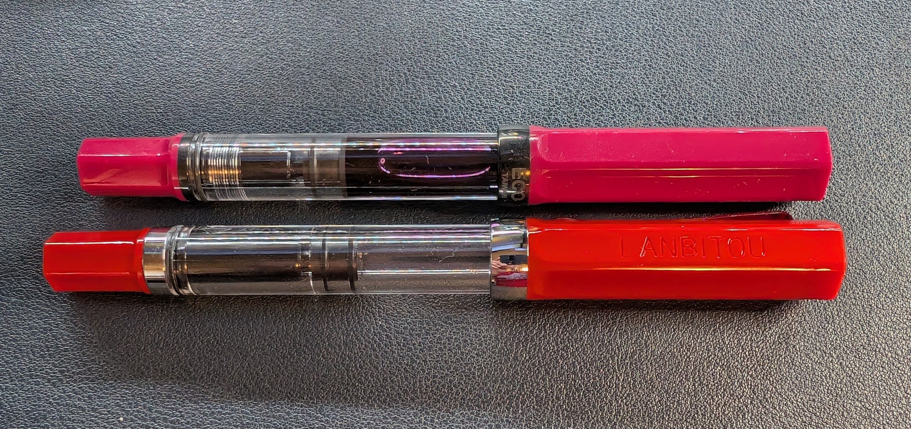Photo of the Lanbitou 3059 pen next to a TWSBI ECO pen. The ECO is on top and has a transparent body with a fuchsia cap and tail/piston knob, with dark metal accents. The 3059 is on the bottom and has a transparent body with a bright red cap and tail/piston knob, with silver accents. It is slightly longer than the ECO and there are subtle differences all over, such as: Metal ring at the base of the piston knob, longer knob, slightly longer cap, different shape of the cap base, different end position of the piston, and different clip shape (not in photo)