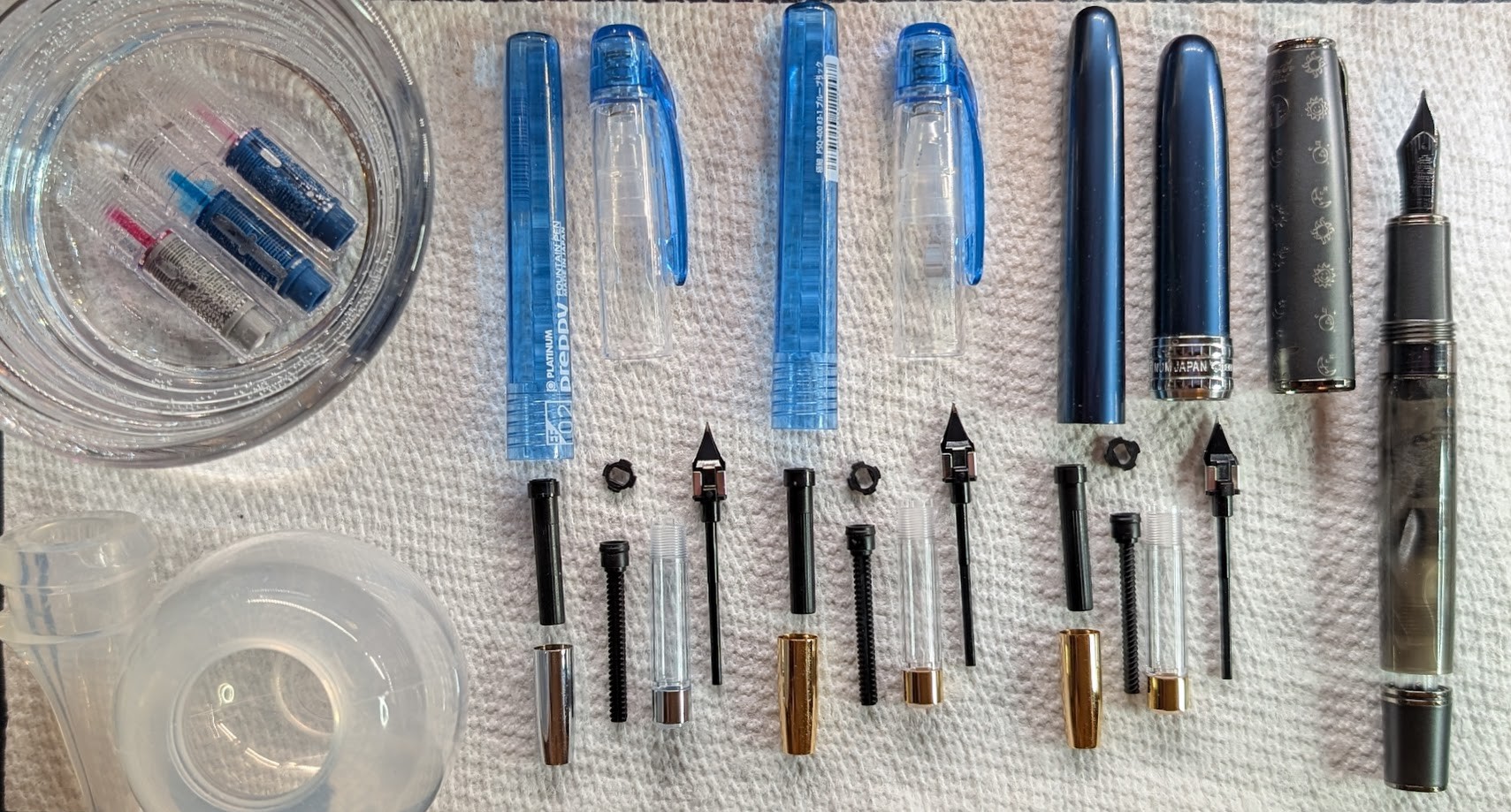Photo of four fountain pens, three of which are disassembled. Plus a glass ramekin full of water in which three of the sections are soaking. From right to left the pens are: Platinum Preppy (transparent blue) x2, Platinum Paisir (blue), and Hongdian N7 Gray Rabbit. All of the Platinum pens are disassembled as much as possible. The sections are soaking in the water in the ramekin. The nib and central part of the feed attached to the nib for each one is below the cap. Next to each nib are parts of the completely disassembled converter. In the lower left corner is a clear bulb syringe with the tip separated.