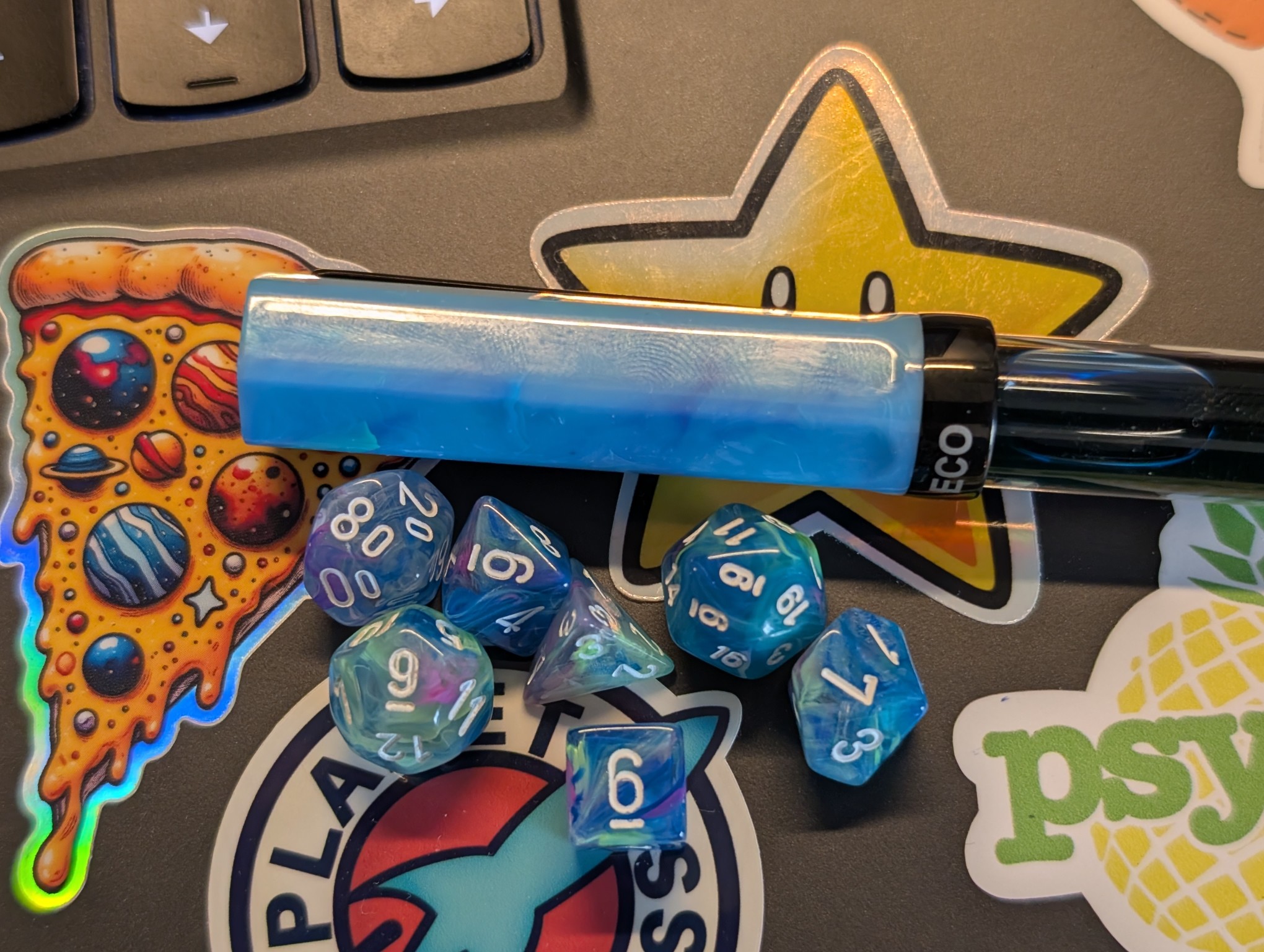 A set of small dice on a laptop. The dice are a marbled mix of blue, green, purple, and clear resin. They are laying next to a fountain pen which is similar in appearance, but mostly blue and not clear in the cap.