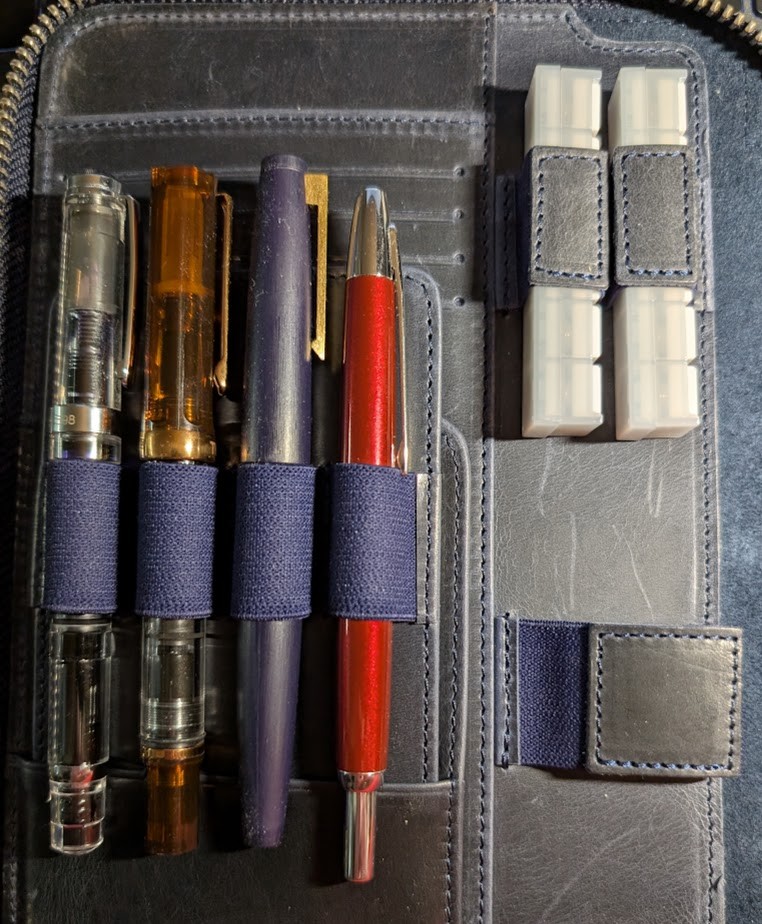 Photo of half the inside of a Galen Leather folio in Crazy Horse Blue. Two stamps are held firmly in loops at the upper right of the photo. Four fountain pens are in other pen loops on the left (A Wing Sung 698 demonstrator, a TWSBI ECO Root Beer w/Bronze, a Jinhao 80 in Blue, and a Pilot VP Decimo in Red)