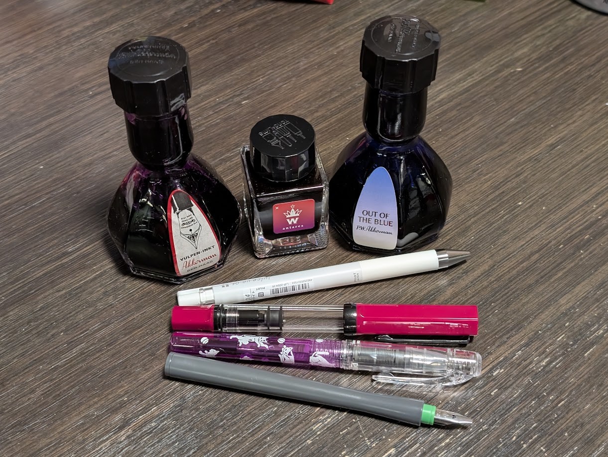 Photo of three ink bottles and four pens. In the back are three ink bottles, two very tall bottles flanking a short bottle. The taller bottles are Akkerman 13 Simplicities Violet, and Akkerman Out of the Blue. The middle bottle is Tono & Lims Antares. Four pens are arranged with their tails touching and tips are spread apart. From top to bottom, they are: - Pilot Juice Up Gel Pen in white with silver trim - TWSBI ECO Plum w/Onyx, it has a bright fuchsia cap and tail and black trim. - Preppy Wa Edo-Monogatari Neko, which is transparent purple plastic with images of white cats playing with balls - Sailor Hocoro 2.0 stub dip pen, with a dark gray barrel
