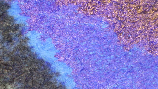 Photo of an ink swatch under magnification. The lower left is black from a stamp on the paper. Next to that is a line of cyan, then a wider strip of purple, then an area that is more red and reflective.