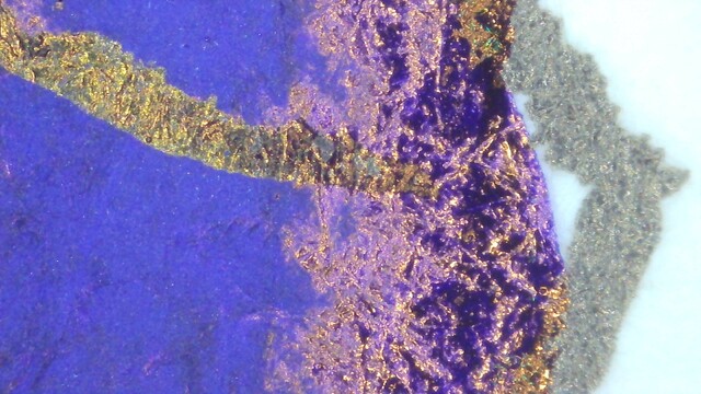Photo of an ink swatch under magnification. There is an outline on the paper visible. The left part is mostly blue, the edge is shiny red with some purple between paper fibers. There is a line of almost gold going through the blue, which is where the ink went over the line stamped on the card.
