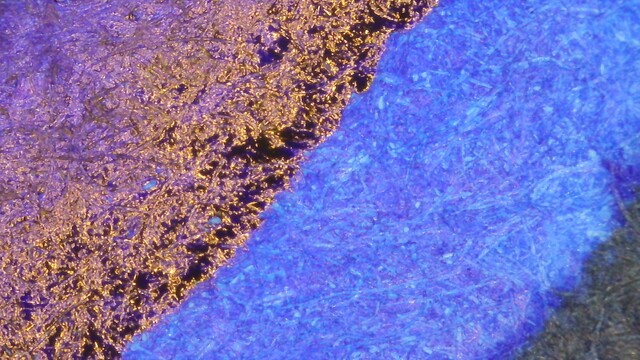 Photo of an ink swatch under magnification. The lower right is blank ink stamped on the paper. After that is a wide line of blurple. Then an area that is red and reflective, and finally a little area of purple in the upper left.