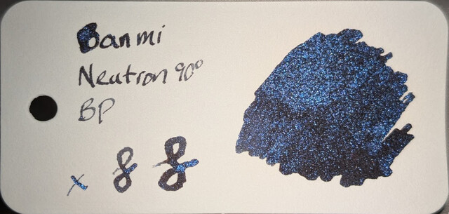Photo of an ink swatch on a col-o-ring swatch card.  Text on the left reads "Banmi Neutron 90° BP" with an "x" and two loops next to it. On the right is a large ink blob made with a glass dip pen.  The base ink is dark, black or extremely dark gray. The ink contains many tiny reflective particles of blue glitter which catch the light.