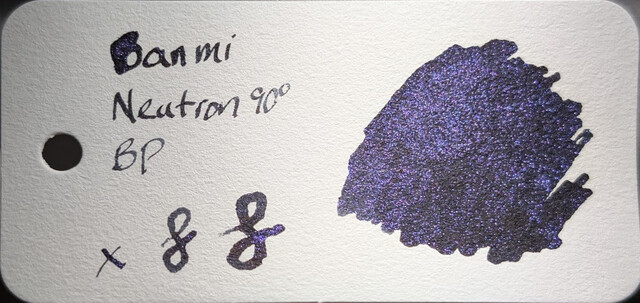 Photo of an ink swatch on a col-o-ring swatch card.  Text on the left reads "Banmi Neutron 90° BP" with an "x" and two loops next to it. On the right is a large ink blob made with a glass dip pen.  The base ink is dark, black or extremely dark gray. The ink contains many tiny reflective particles of purple glitter which catch the light.