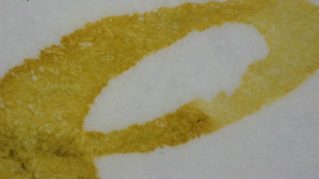 Photo of ink lines on Midori paper under magnification. The ink is dark yellow-gray, with some areas lighter and other areas darker, especially in a couple small places where paper fibers appear very dark yellow. The edges of the lines are somewhat rough.