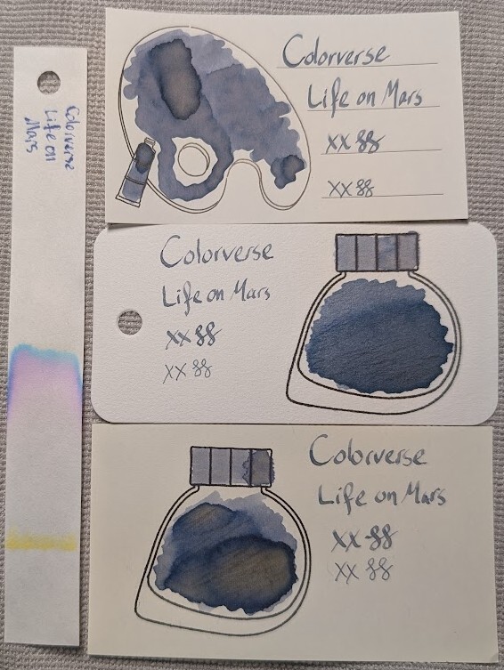 Photo of 3 ink swatches & chromatography strip for Colorverse Life on Mars which is a "dusty" blue ink with chromashading in swatches and some shading in text. Each swatch contains the ink name followed by "XX" 2 curlicues in both stub and EF widths. Swatch 1: Iroful swatch card with an outline of a paint palette and tube. The palette is mostly filled in with swirls of ink, a few small areas are darker a lighter gray, almost brown center. There is an outline of a paint tube which is swabbed and looks similar. The writing has a lots bit of shading across letters with wider lines. Swatch 2: Col-o-ring swatch card with a stamped outline of a Colorverse ink bottle, which is teardrop-shaped with the point at the bottom left. The outlined area is mostly full of an ink blob which is darker and more gray at the bottom and paler and more blue at the top in a gradient. Swab across "cap" is similar, but more gray. Again, the words in the sample have a some shading. Swatch 3: Midori MD swatch card with a stamped outline of a Colorverse ink bottle. The outlined area is mostly filled in with swirls of ink, a few areas are darker with even darker edges and gray interiors. Swab across "cap" is similar. The text has much more pronounced shading and on nearly every letter. Chromatography is visible on most of the strip, there is a line of yellow where it started. A large faint area of blue. Then blends into pink, then purple, then light cyan.