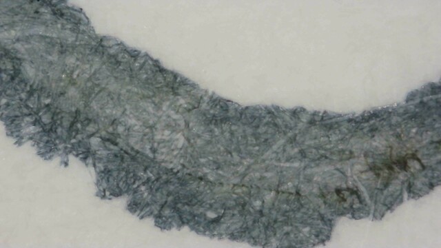 Photo of ink writing on Col-o-ring paper under magnification. The ink is blue-gray, slightly darker at the edges, and blue-green near the center. There are some small dark paper fibers which are almost black near the bottom center of the line. The edges of the line are very rough.