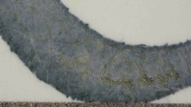 Photo of ink writing on Iroful paper under magnification. The ink is blue-gray, darker and more blue at the edges, some areas around the middle are yellow-ish in the background. Some paper fibers near the center are much darker, almost black. The edges of the line are smooth.