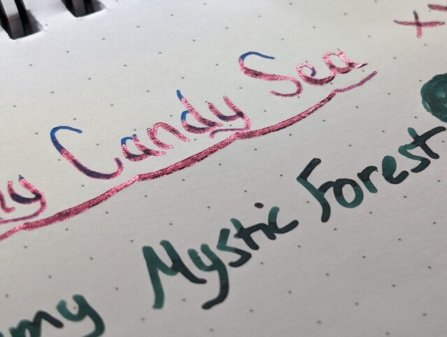 Writing sample on dot grid paper  First line is partially visible, reads "y Candy Sea" followed by an "x". The text is underlined with a variable-width line. The ink is blue and there is a noticeable reddish sheen on portions of the lines in the writing.  Second line is partially visible, reads "my Mystic Forest" followed by a green blob. The ink is dark green.
