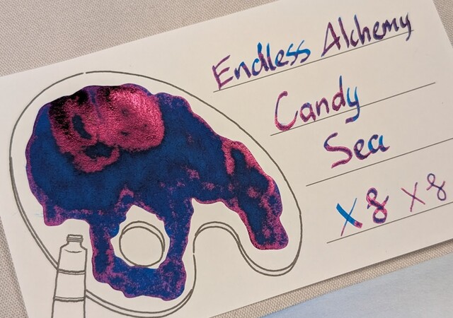 Low angle photo an ink swatch card.  Overall the ink is a deep, dark blue with heavy dark red reflective sheen.  The card is an Iroful swatch card with an outline of a paint palette. Inside the palette is mostly filled in with swirls of midnight blue ink and areas reflecting magenta. Some areas with more ink are even darker blue, and areas with even more ink are reflecting magenta.